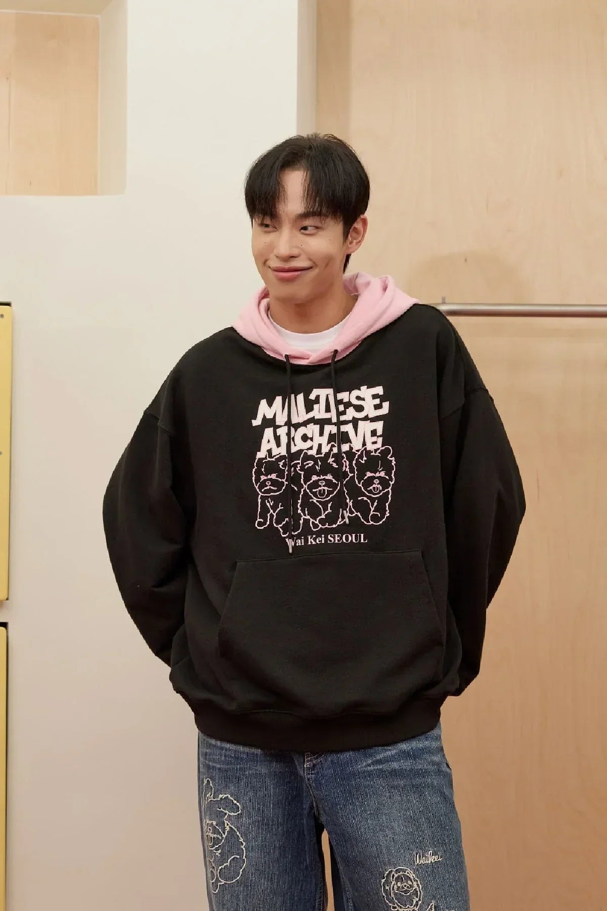 Man wearing black Maltese Archive hoodie with pink hood and graphic dogs, paired with denim jeans with dog embroidery