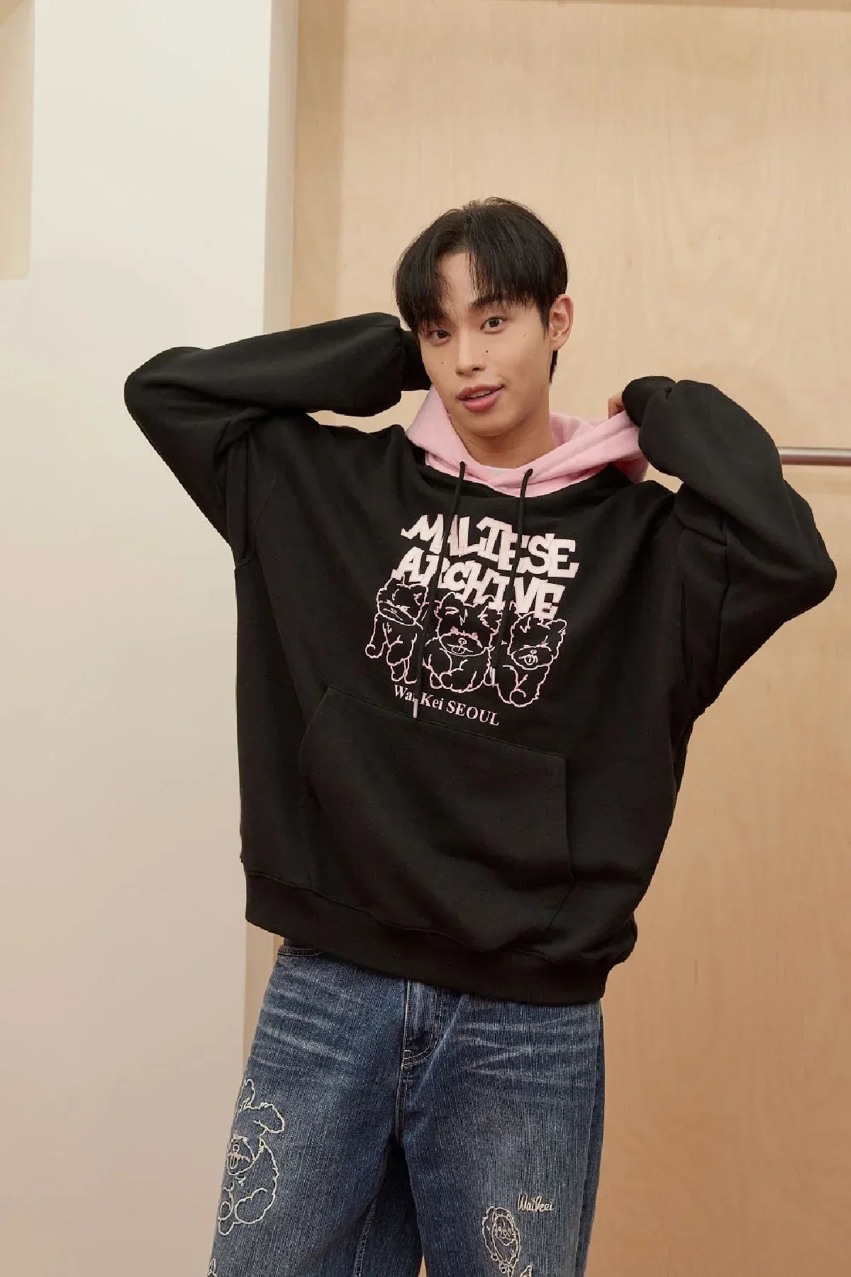 Young man wearing black Maltese Archive graphic hoodie with pink hood and embroidered blue jeans indoors