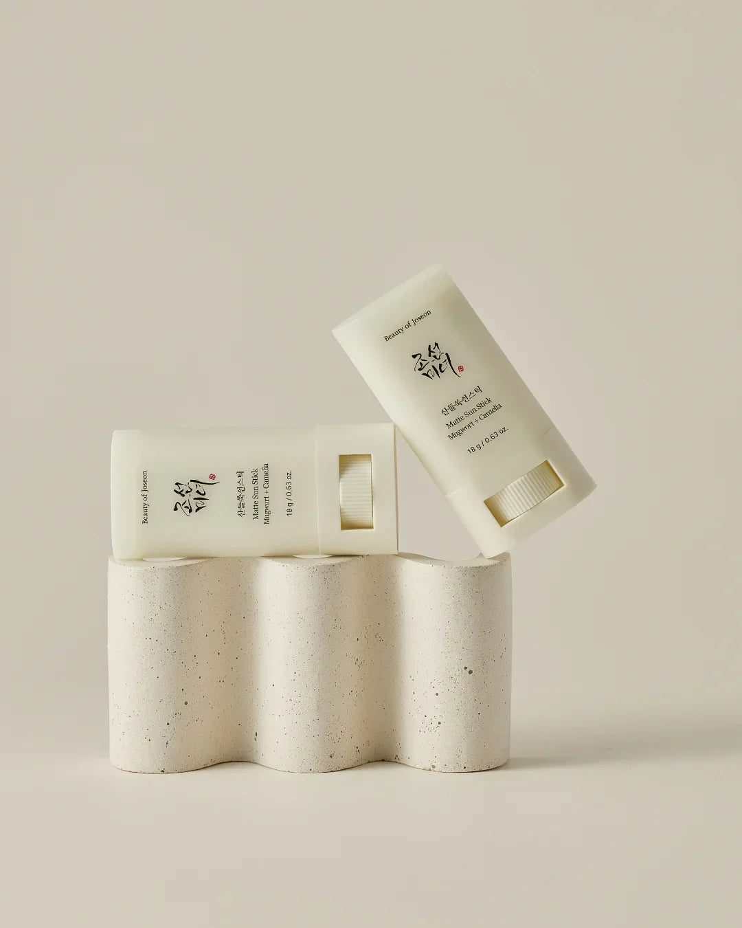 Two matte sun stick containers with Mugwort and Camellia, placed on a beige textured stand