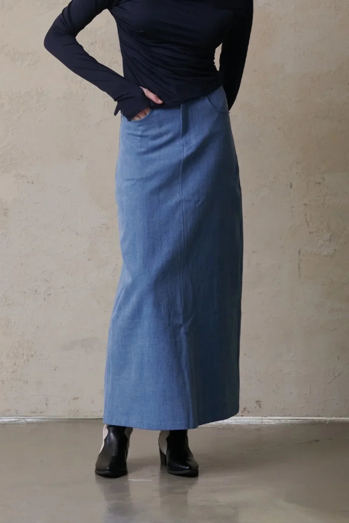 Woman wearing a light blue maxi denim skirt with pockets, black long sleeve top, and black ankle boots