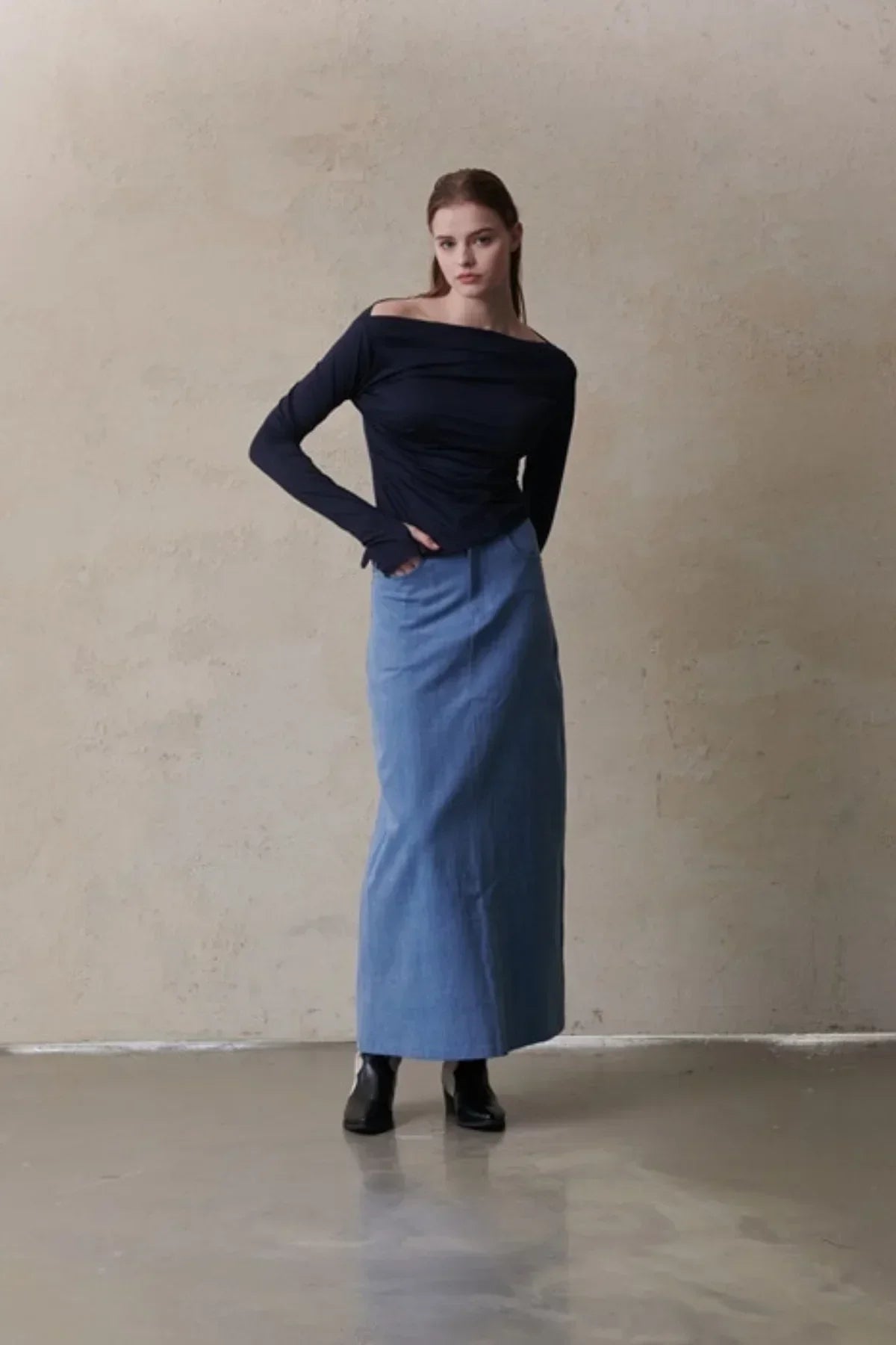 Model wearing a mid-blue maxi denim skirt with a navy off-shoulder long sleeve top and black ankle boots