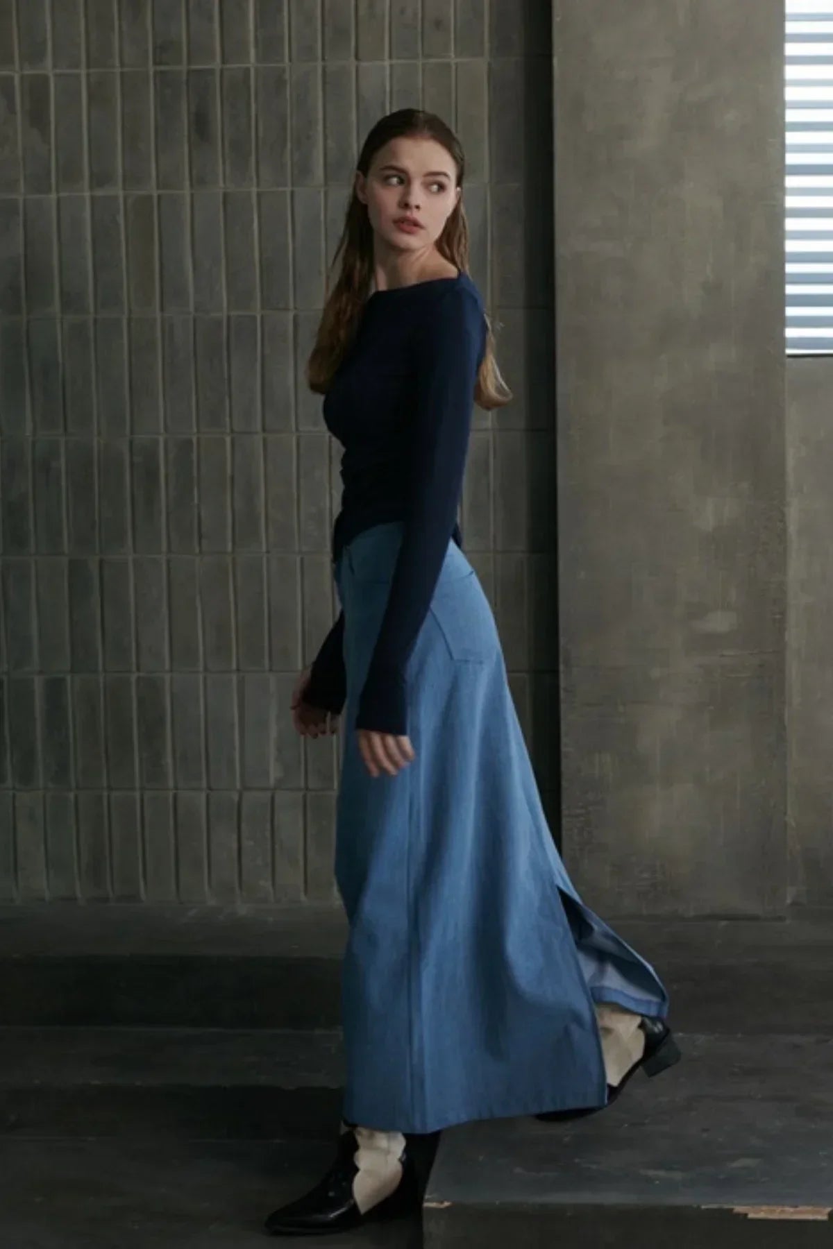 Woman wearing a mid-blue maxi denim skirt with side slit, navy long-sleeve top, and black boots in industrial setting