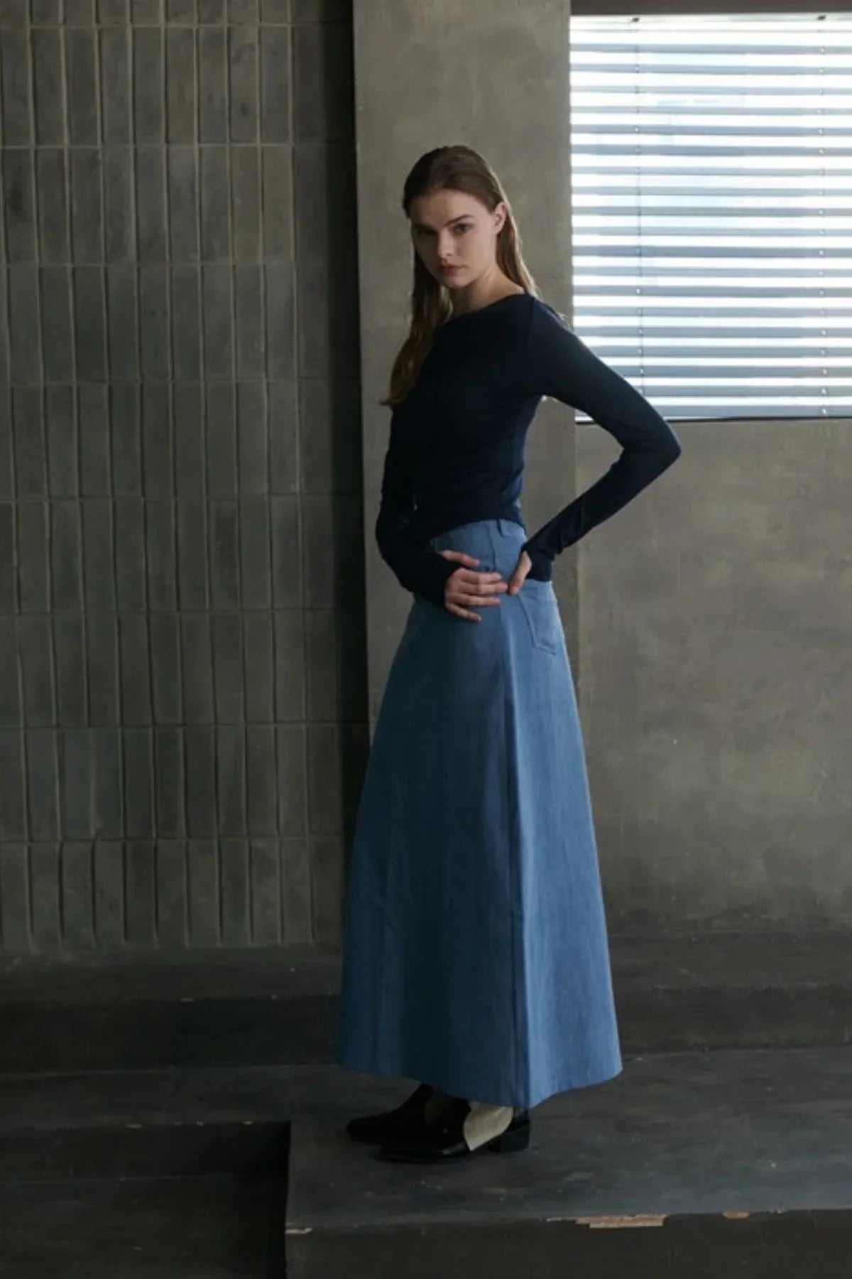 Woman wearing a mid-blue maxi denim skirt with black boots and a long-sleeve navy top in a modern industrial room