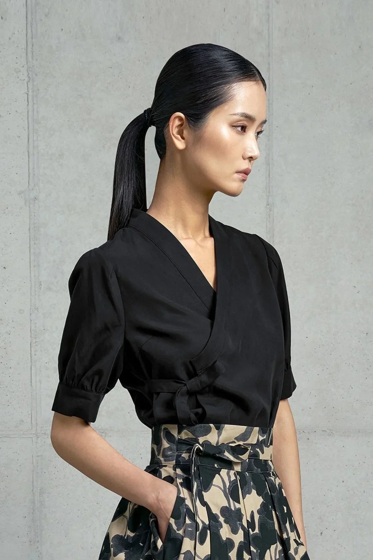 Side profile of woman in black wrap blouse with short puff sleeves and floral high-waist skirt against concrete wall
