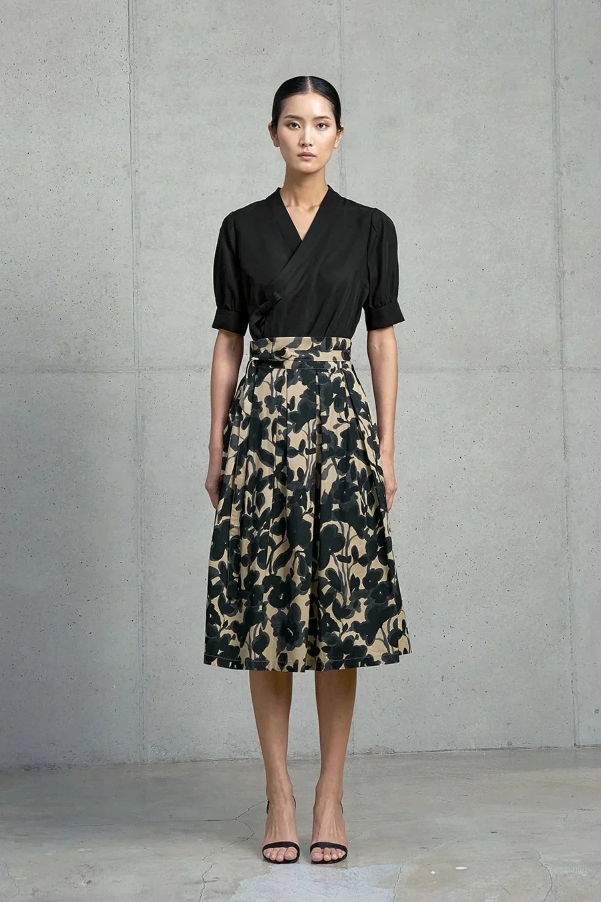Woman wearing black wrap blouse and high-waisted beige skirt with black floral print standing against concrete wall
