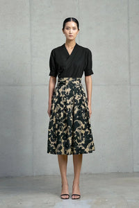 Woman wearing black wrap blouse and high-waisted beige skirt with black floral print standing against concrete wall