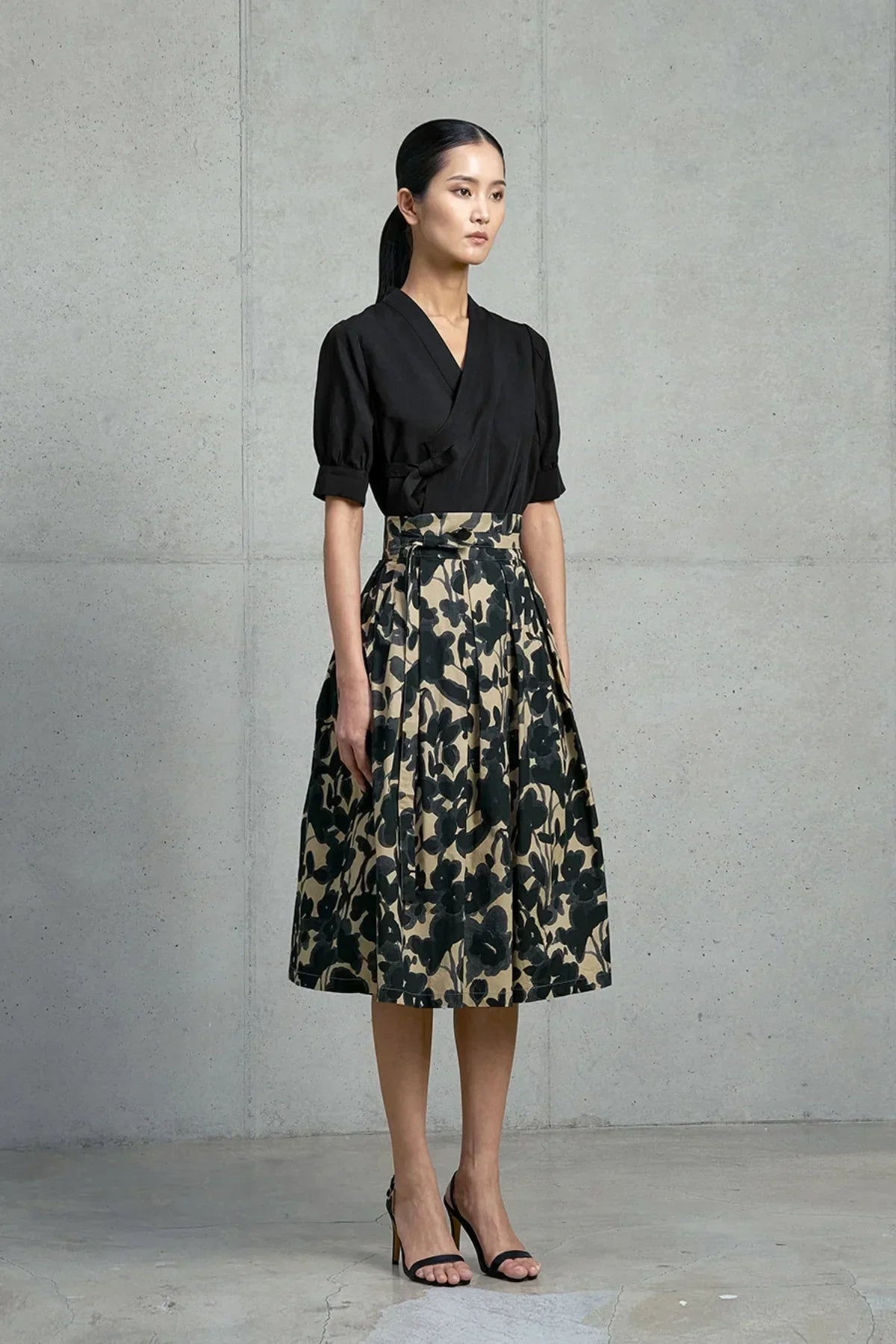 Model wearing black short-sleeve blouse and beige midi skirt with black floral print in concrete room