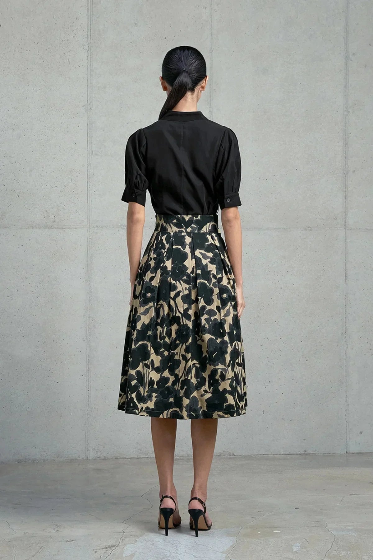 Back view of a woman wearing a black blouse with puffed sleeves and a high-waisted beige skirt with black floral print and black high heels