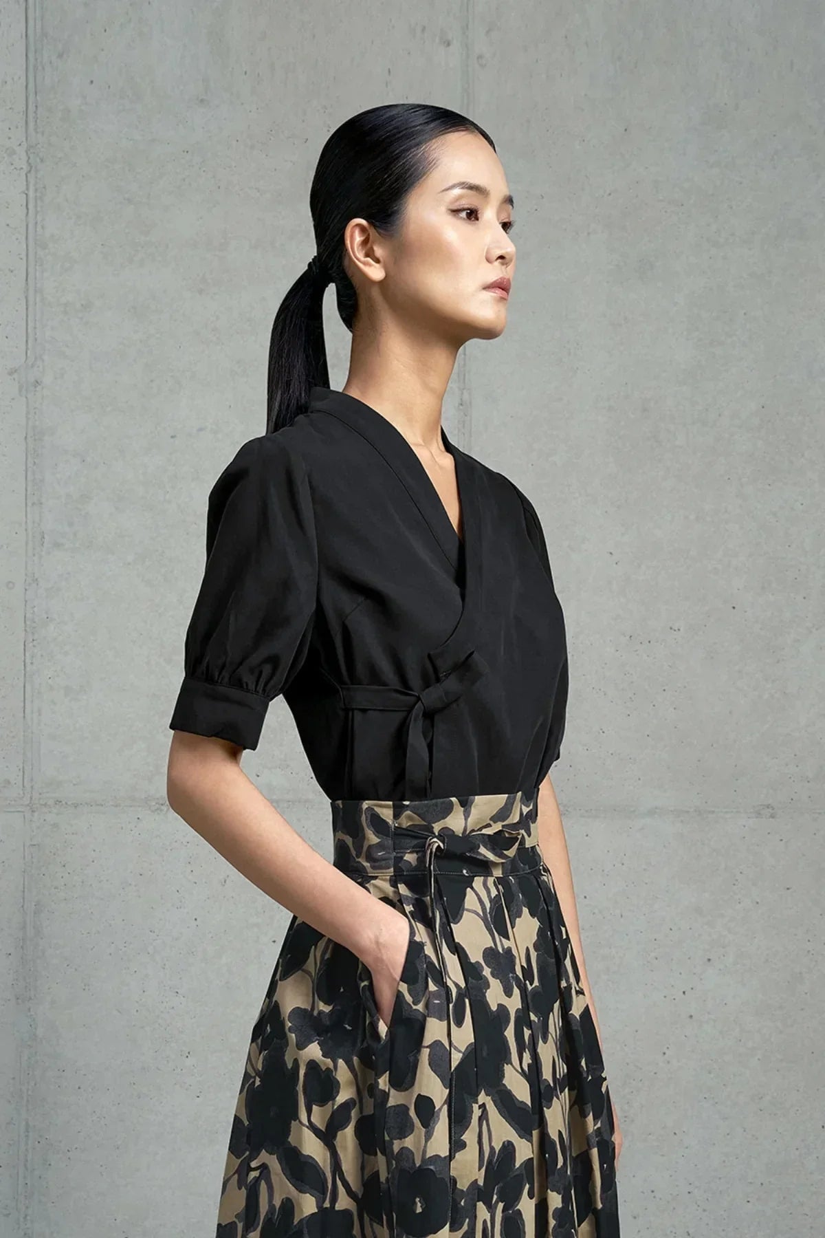 Woman in black wrap blouse and high-waist beige skirt with black floral print against gray concrete wall