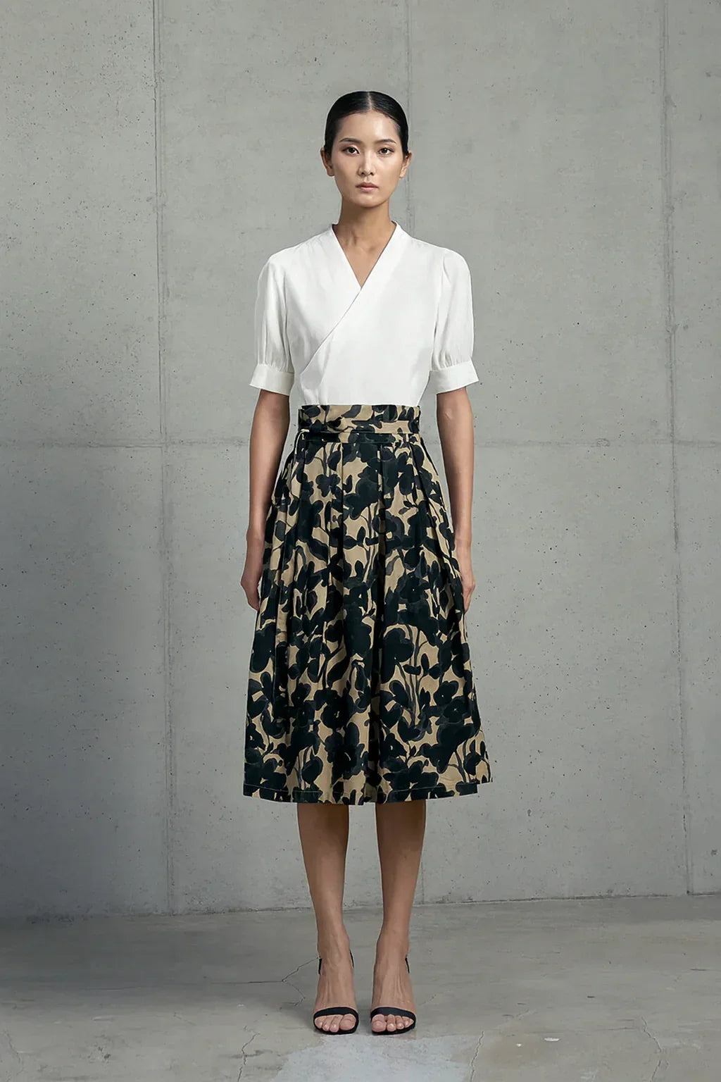 Woman wearing ivory wrap blouse and mid-length skirt with black floral print, standing against concrete wall