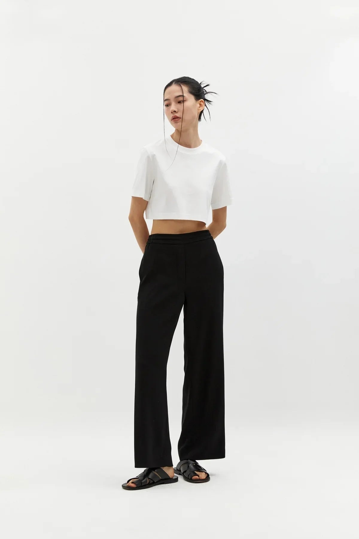 Woman wearing black semi-wide pants and white cropped t-shirt with black sandals on white background