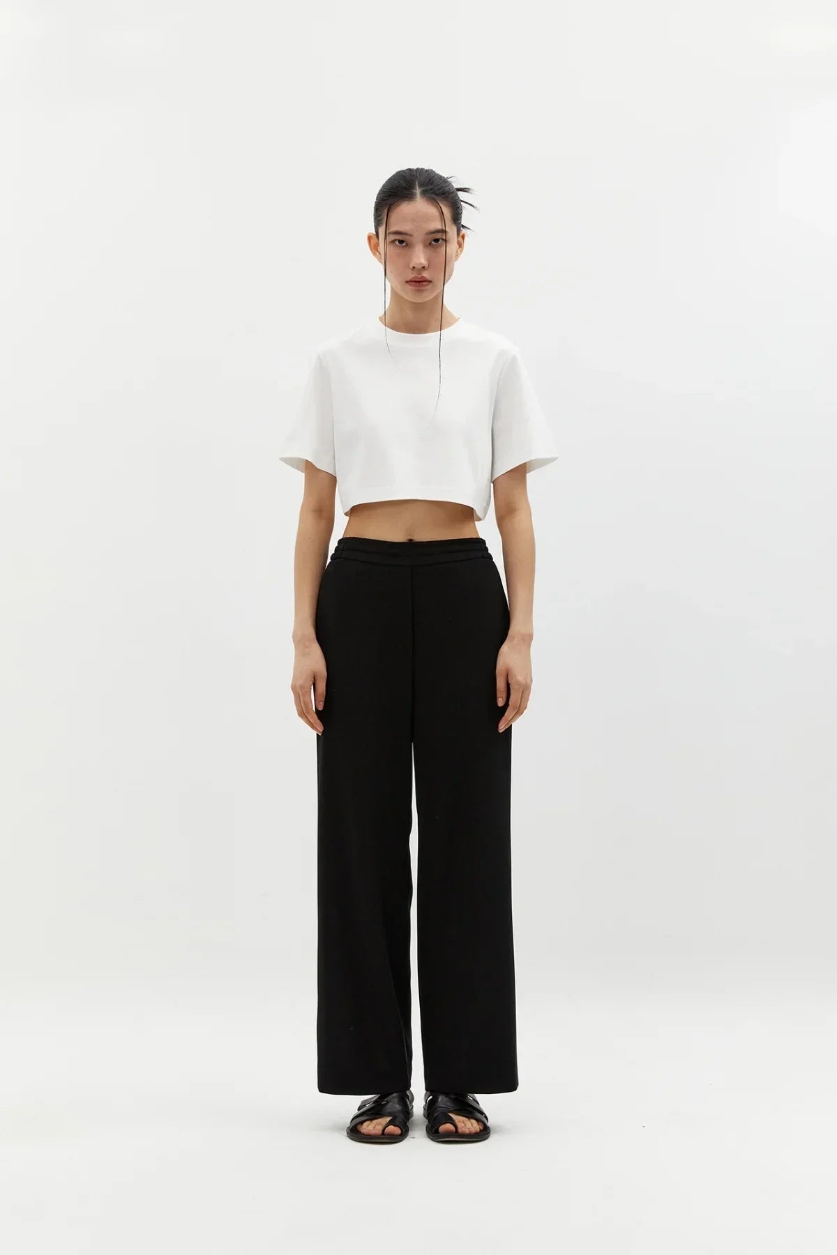 Woman wearing black semi-wide pants with a white cropped t-shirt and black sandals in a studio