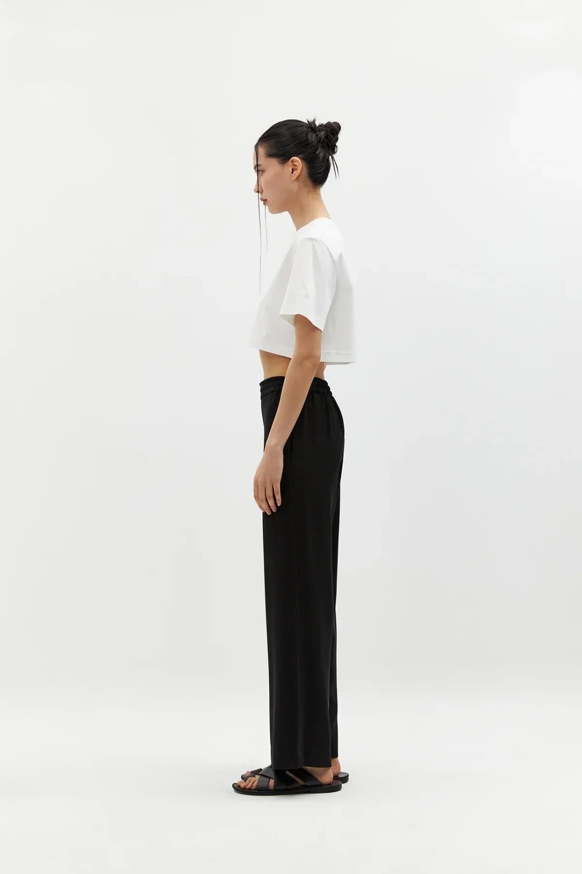 Side view of woman wearing black semi-wide pants and white cropped t-shirt on white background
