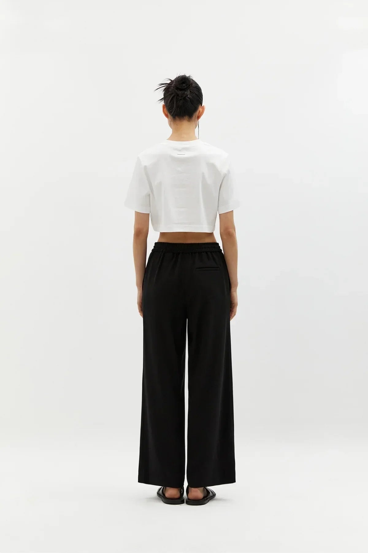 Back view of woman wearing black wide-leg pants and white cropped t-shirt on white background