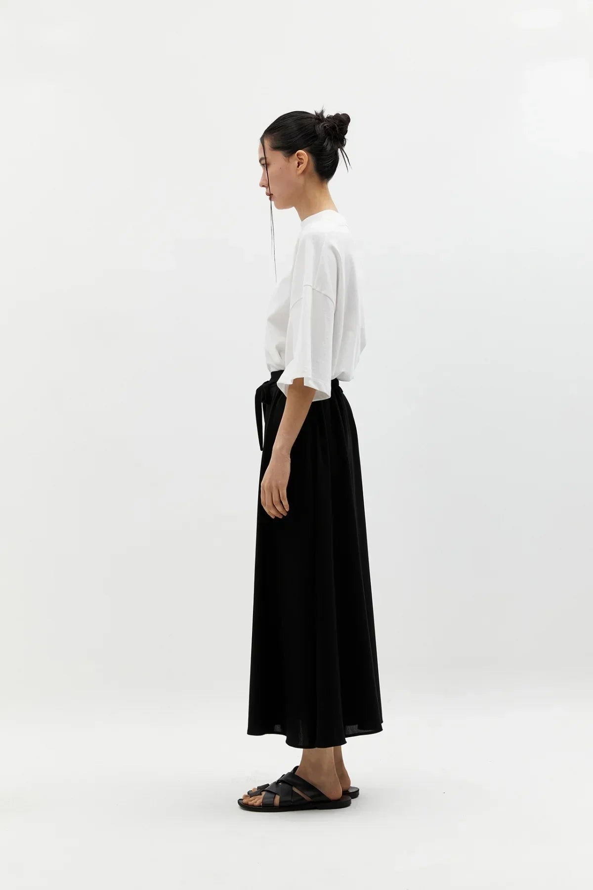 Side view of woman in white oversized t-shirt, black wide skirt with belt, and black sandals on white background