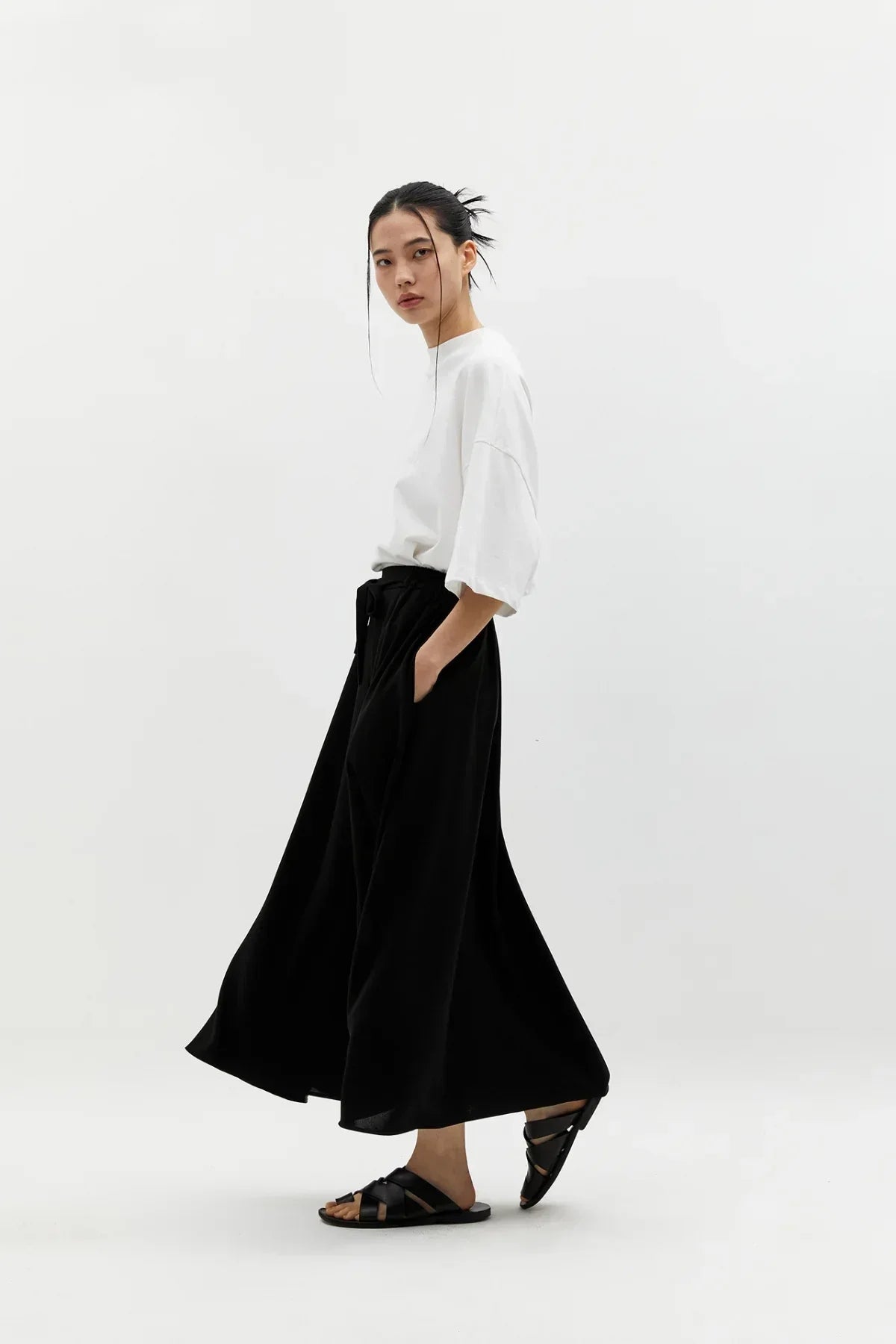 Woman in a white oversized top and flowing black wide skirt with pockets, black sandals, studio shot