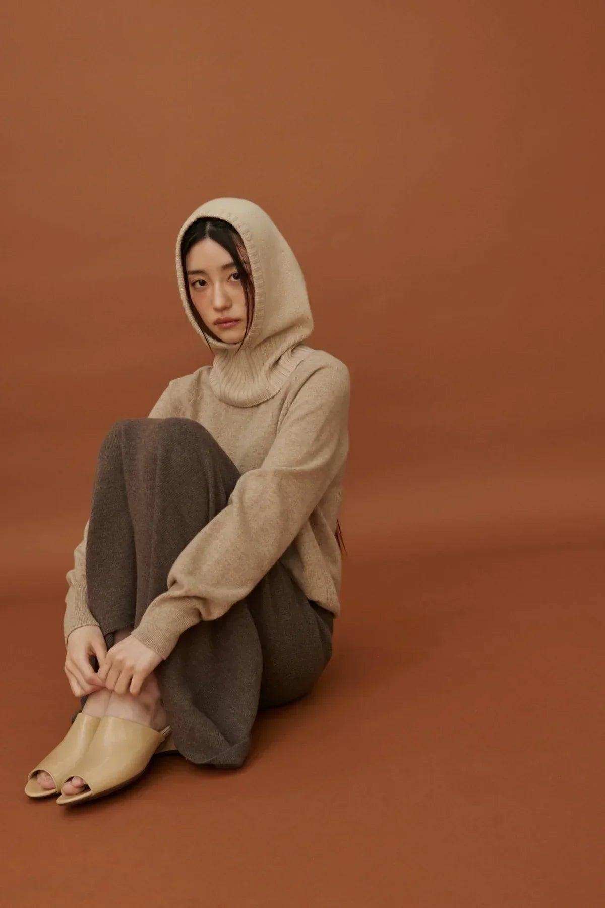 Woman wearing beige cashmere balaclava and sweater with brown pants seated on brown background