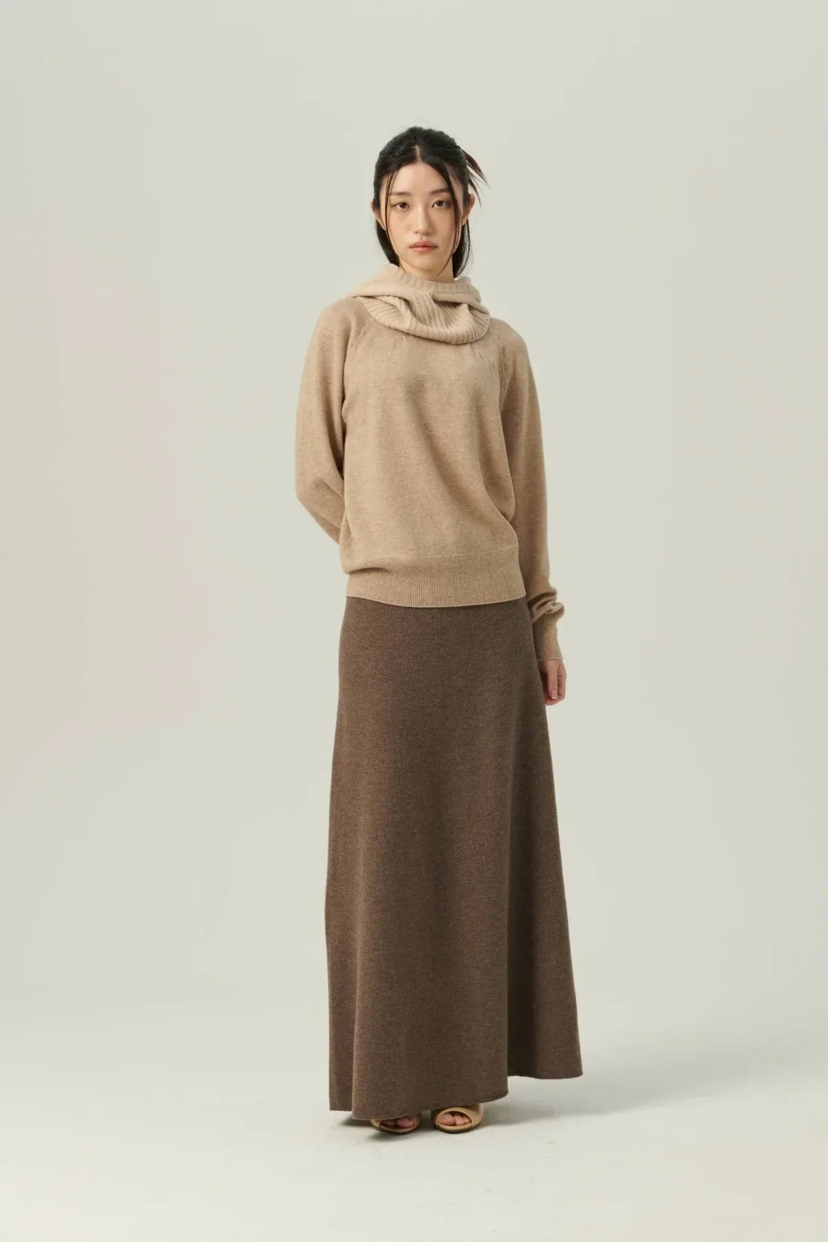 Woman wearing beige cashmere sweater and long brown knit skirt against plain background