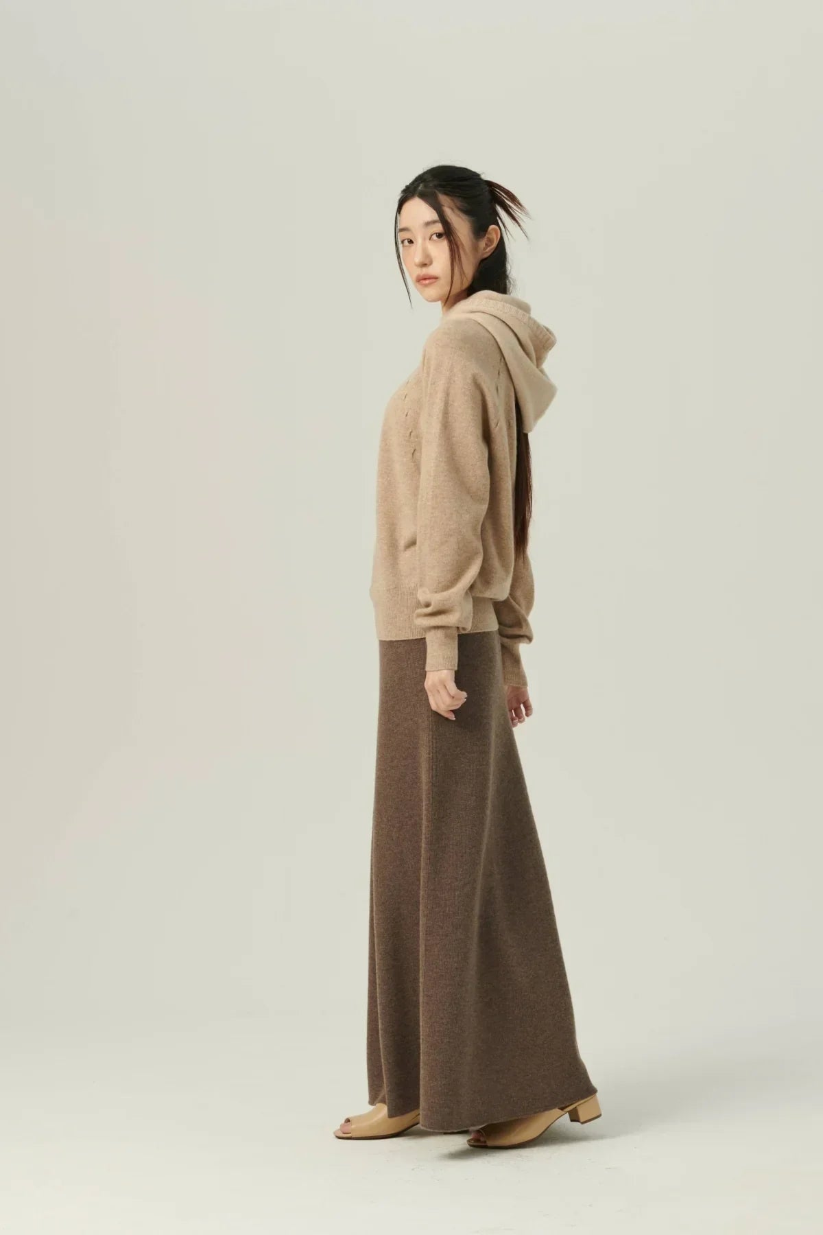 Woman wearing beige wool hoodie and brown maxi skirt with beige open-toe heels on plain background