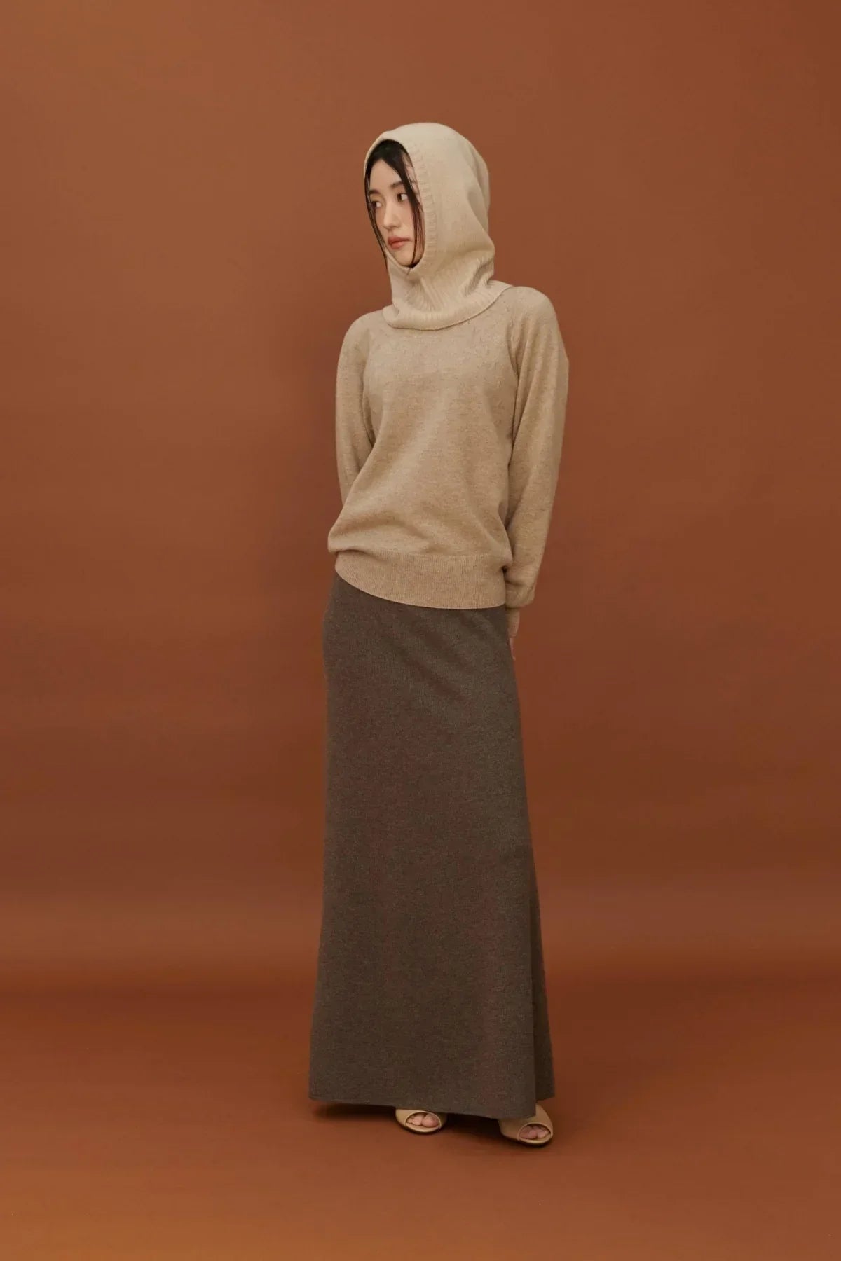 Woman wearing beige cashmere balaclava sweater and dark brown long skirt on brown backdrop