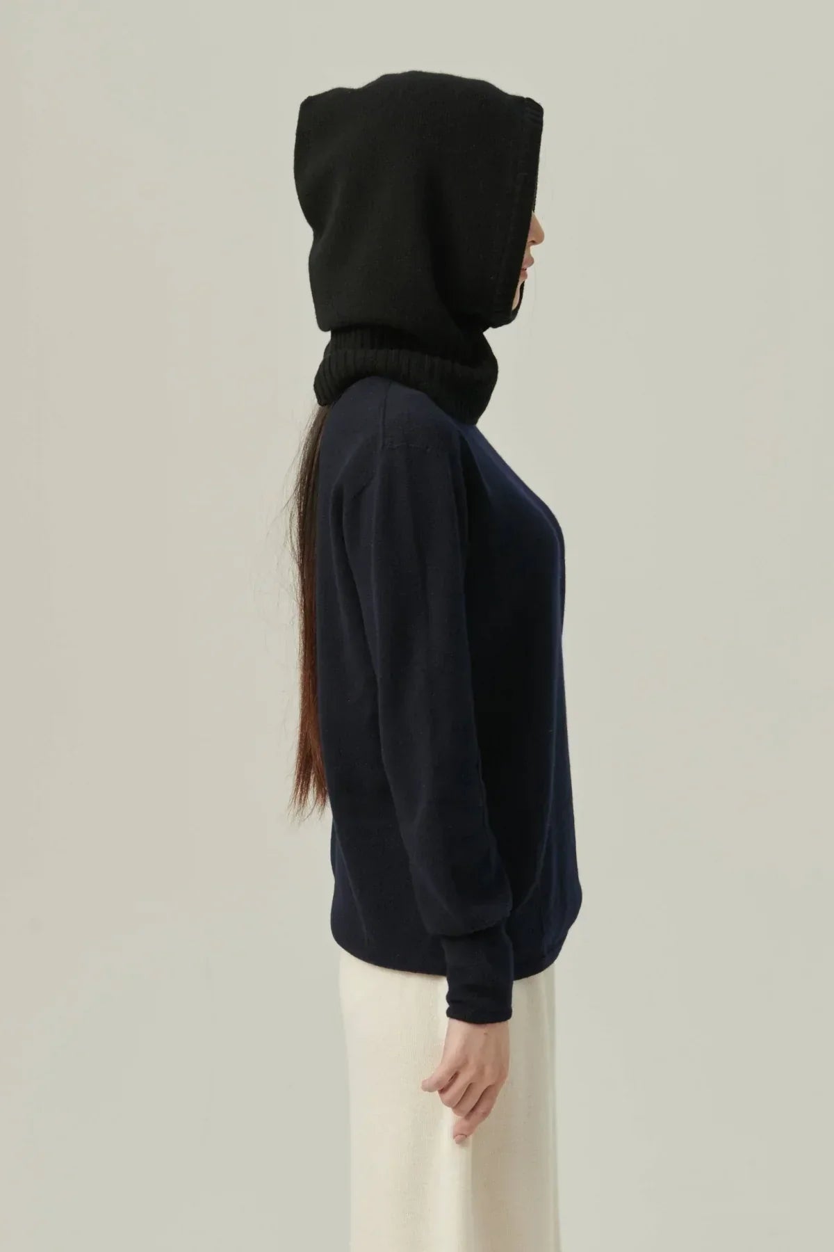 Side view of woman wearing a black cashmere balaclava hood and navy blue sweater against neutral background