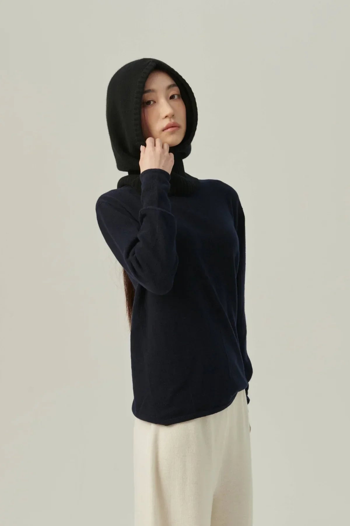 Model wearing a black minimal cashmere balaclava with a navy sweater and white pants in studio