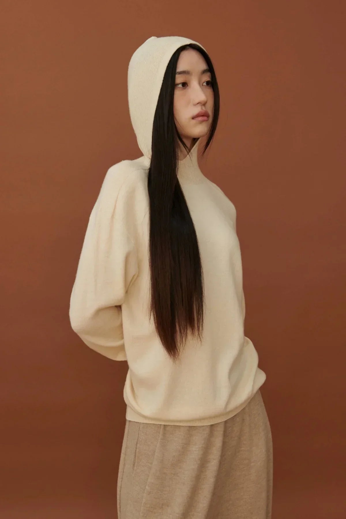Woman wearing cream mock neck hooded knit sweater and beige pants against brown background