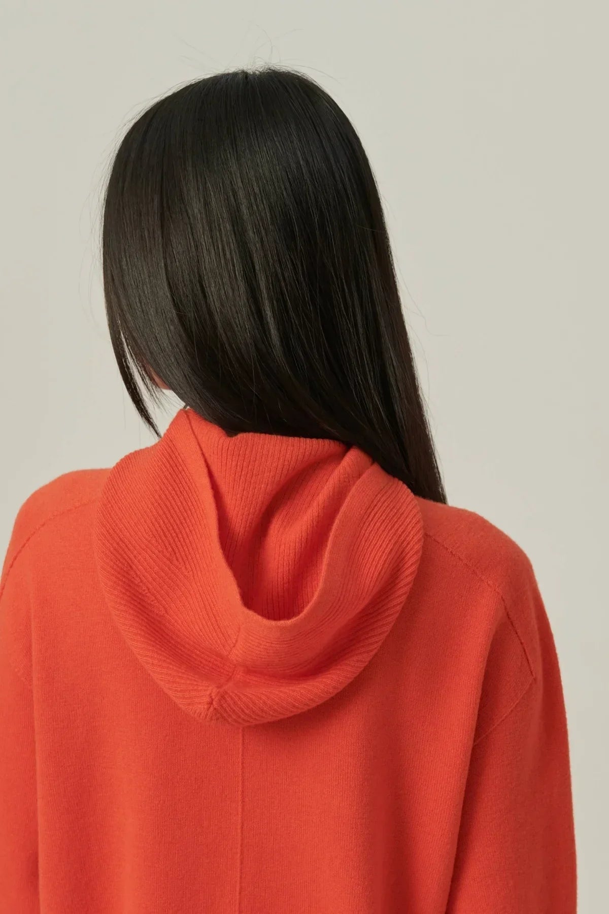 Back view of person with long dark hair wearing an orange hooded knit sweater against plain background