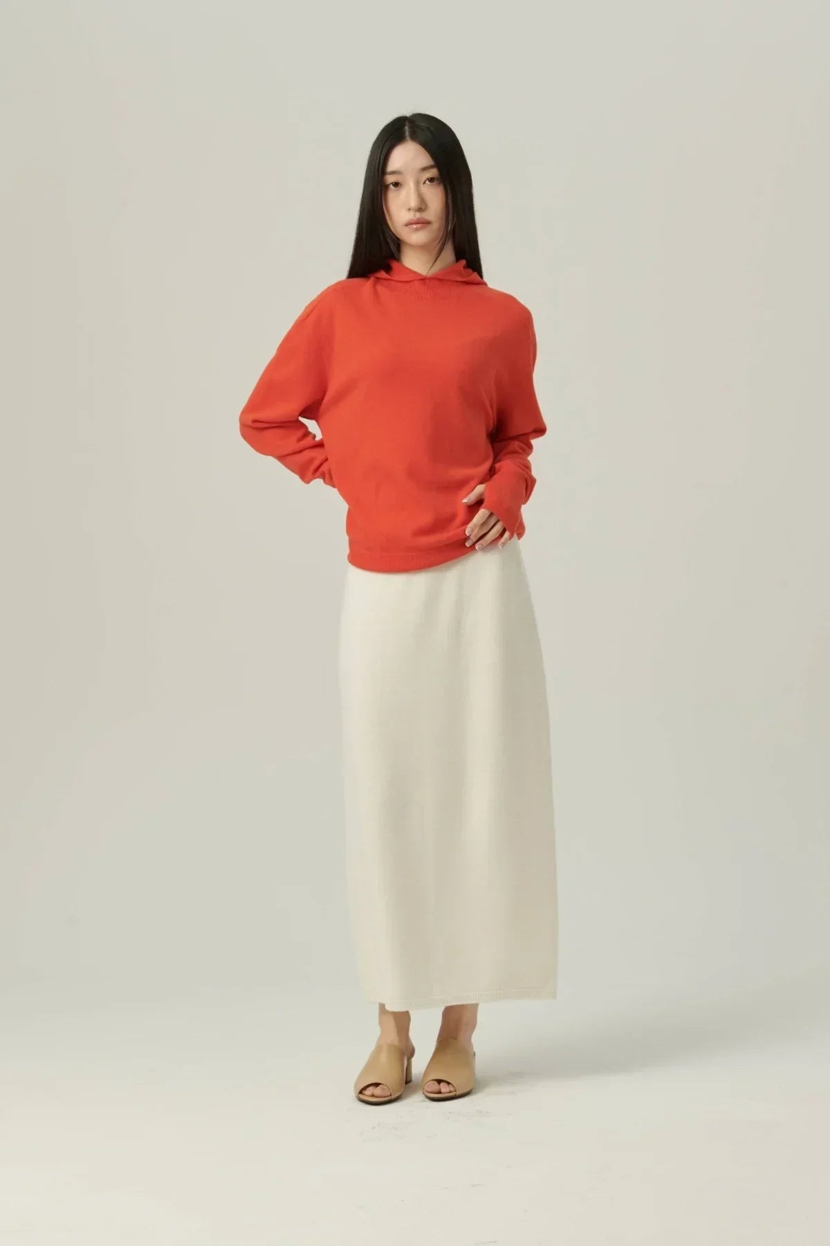 Woman wearing orange mock-neck hooded knit sweater with long white knit skirt and beige open-toe mules