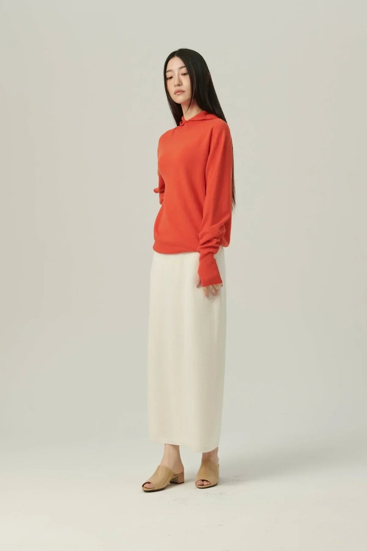 Woman wearing orange mock-neck hooded knit sweater and long cream skirt with beige open-toe shoes