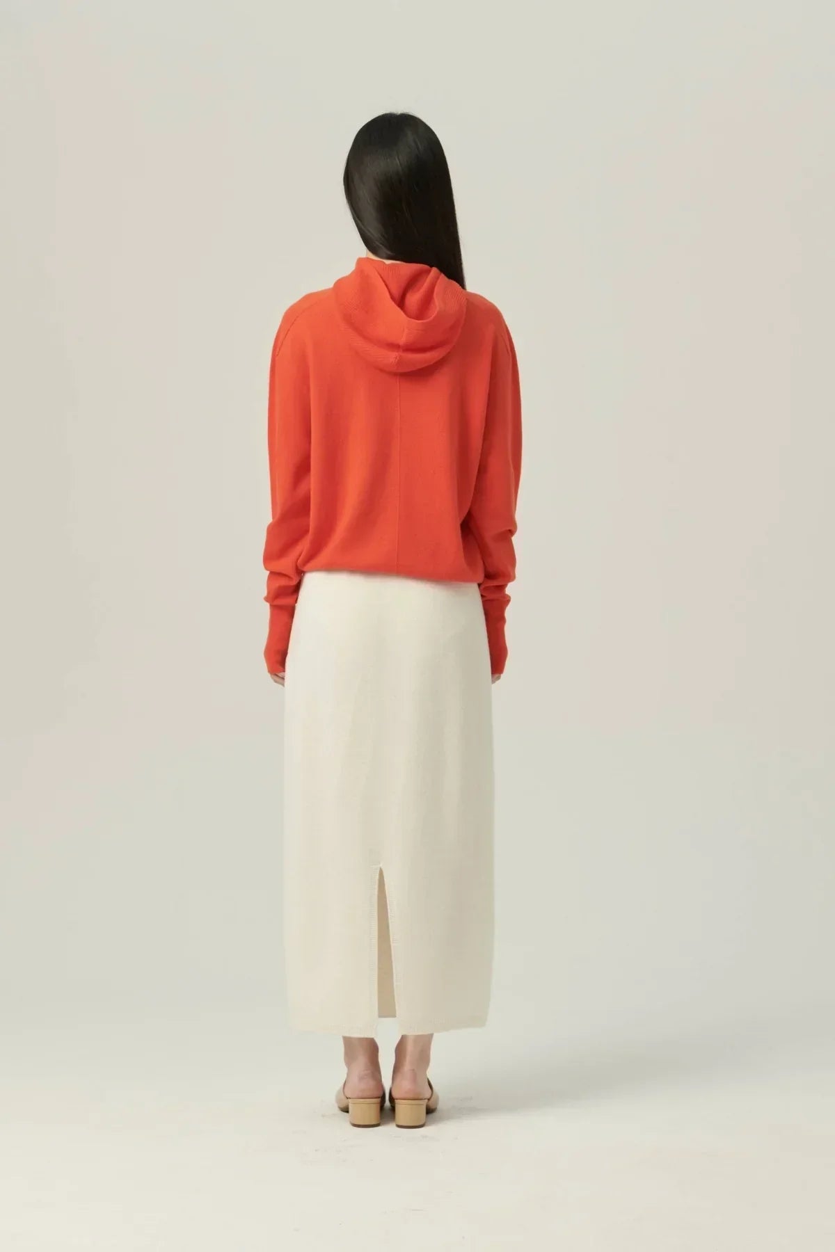 Woman wearing orange hooded knit sweater and cream midi skirt with back slit in studio