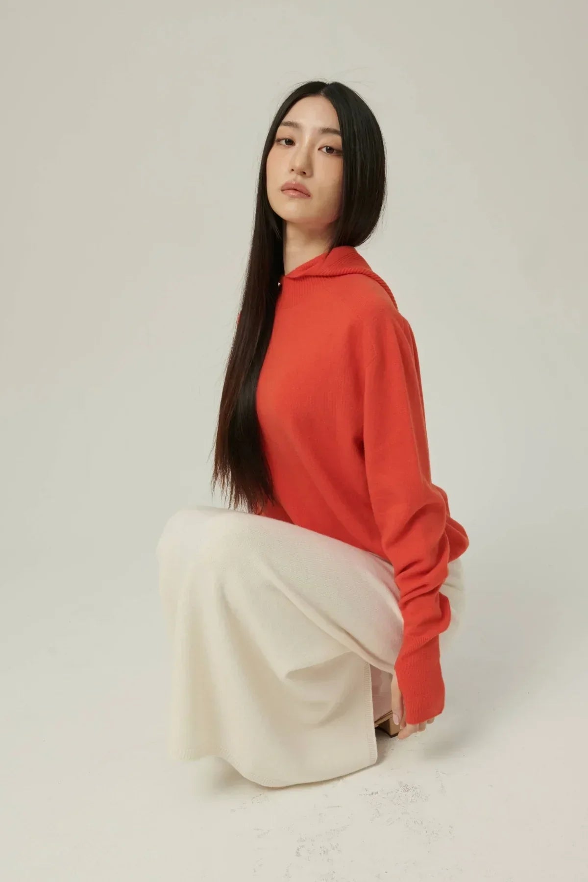 Woman with long black hair wearing an orange mock-neck hooded knit sweater and white skirt, studio background