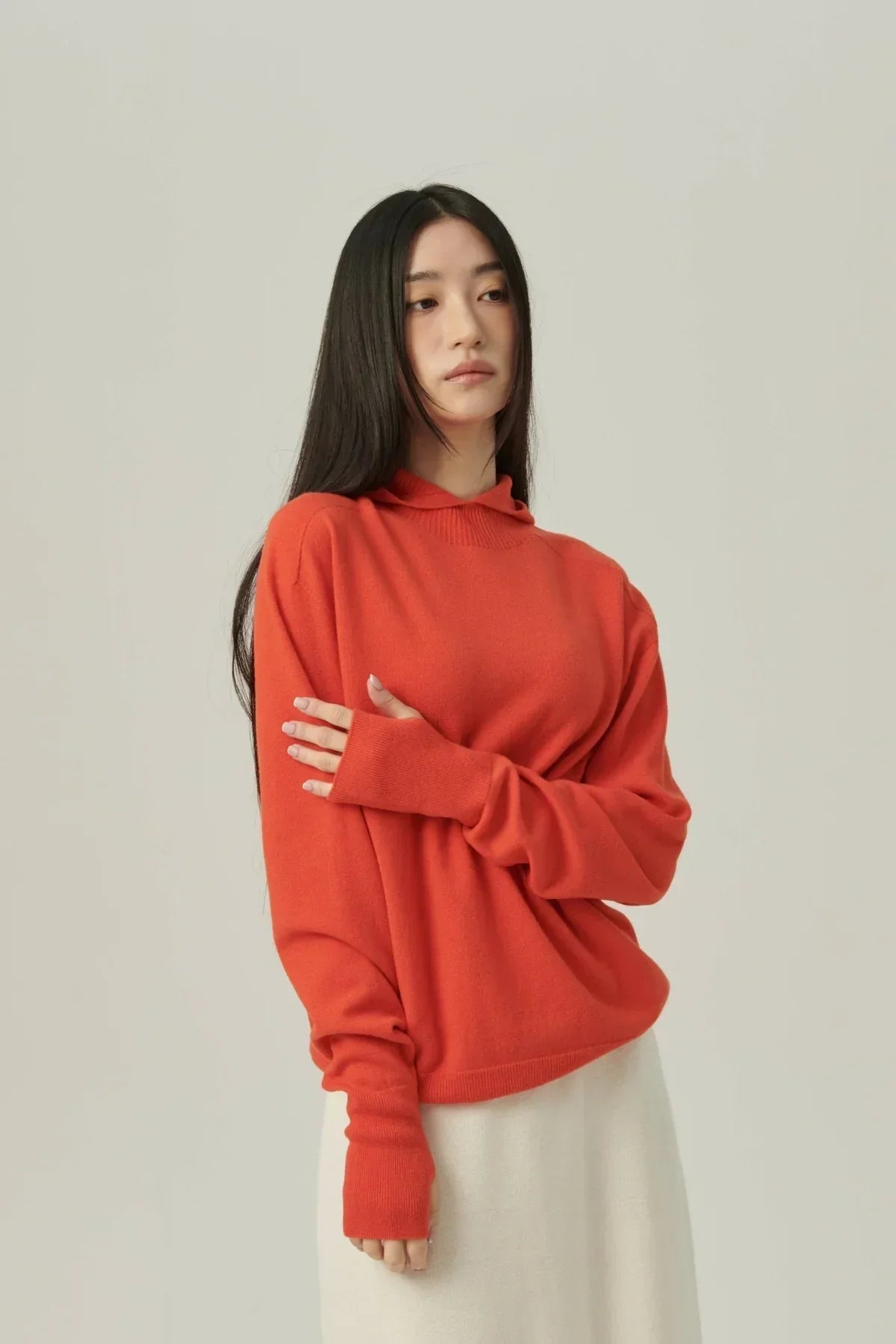 Woman wearing orange mock-neck hooded knit sweater with long sleeves and cream skirt on neutral background