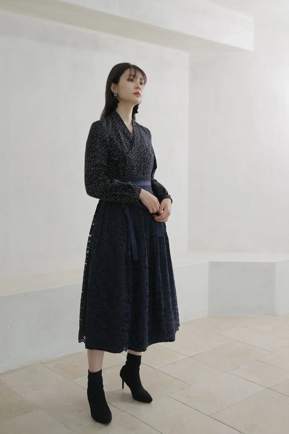 Woman wearing a black floral blouse and long lace skirt with ankle boots in a minimalist light room
