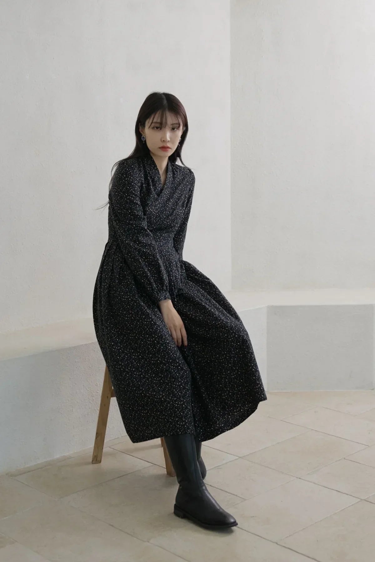 Woman sitting indoors wearing a long black floral dress and black knee-high boots on beige tiled floor