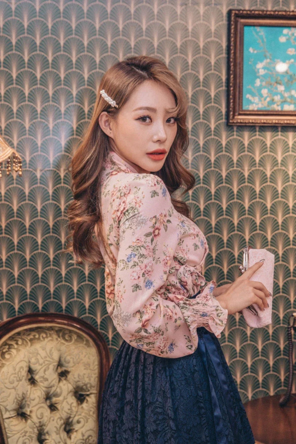 Woman in floral pink blouse and navy lace skirt holding pink clutch in vintage room
