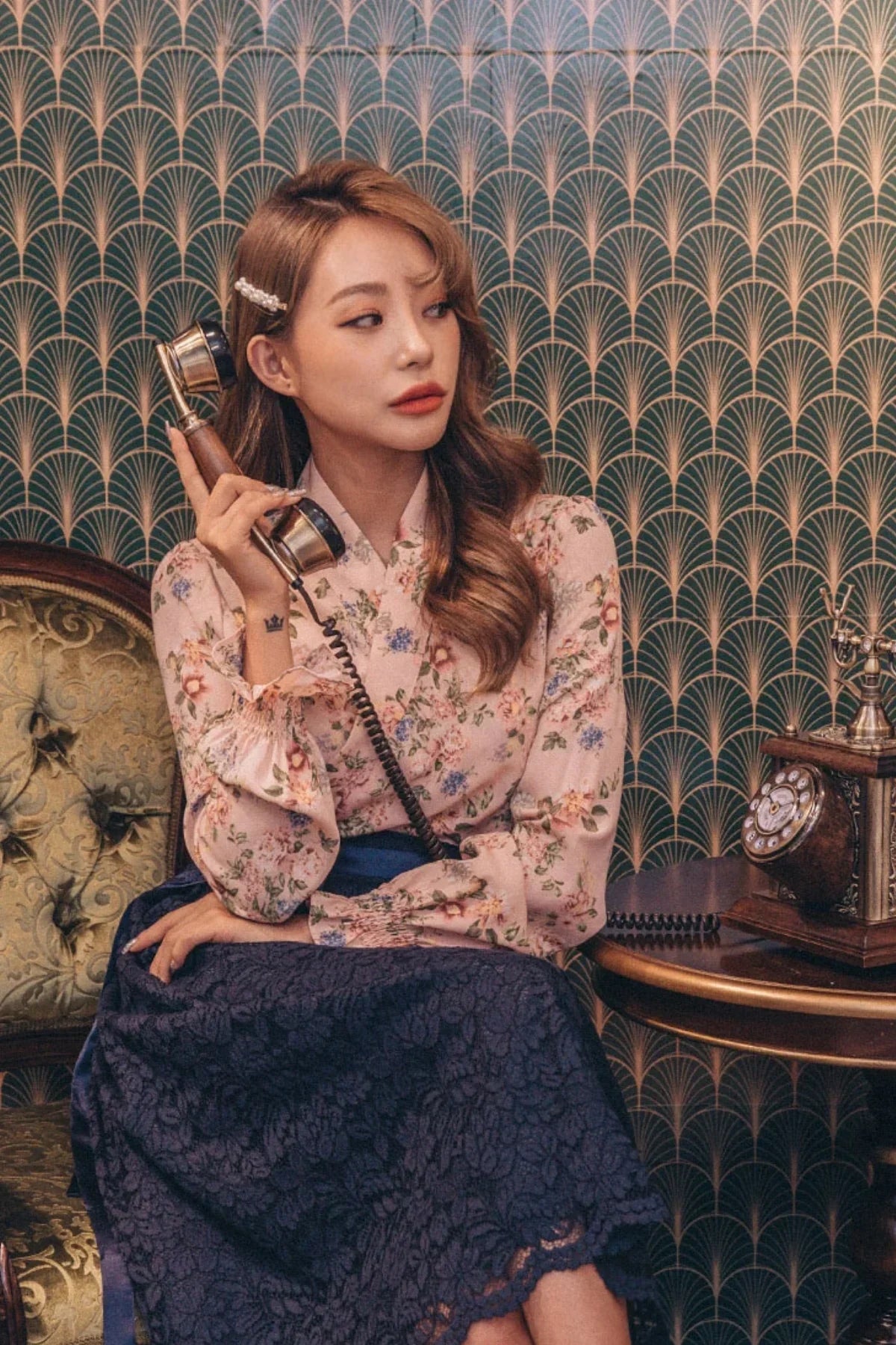 Woman in floral blouse and navy lace skirt sitting on vintage chair holding antique phone receiver