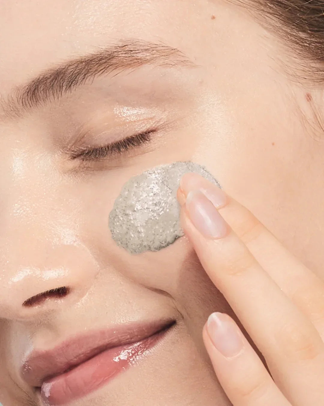Close-up of a woman applying gray mugwort calming clay mask on cheek for skincare