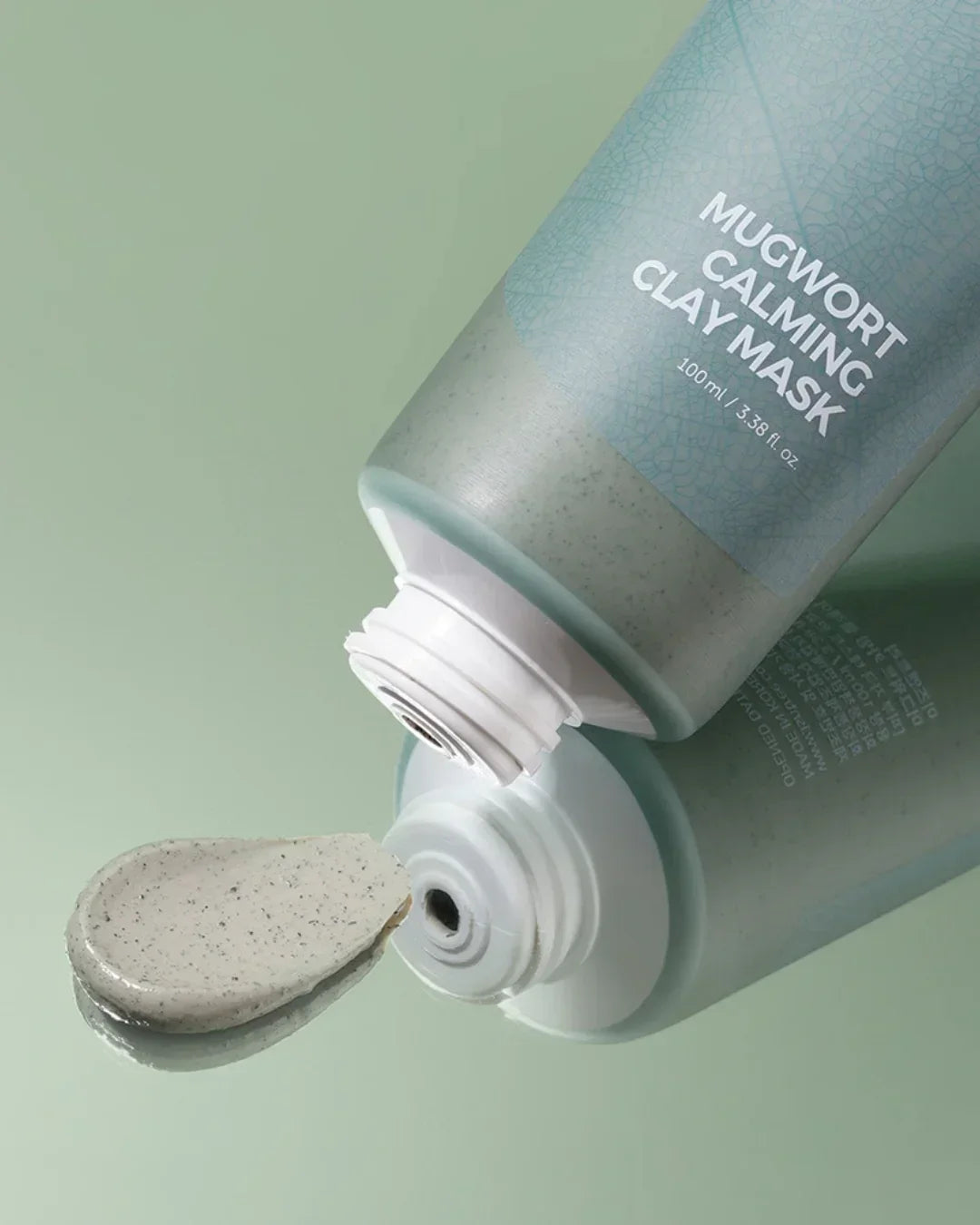 Tube of Mugwort Calming Clay Mask with a dollop of grey speckled clay mask on green surface