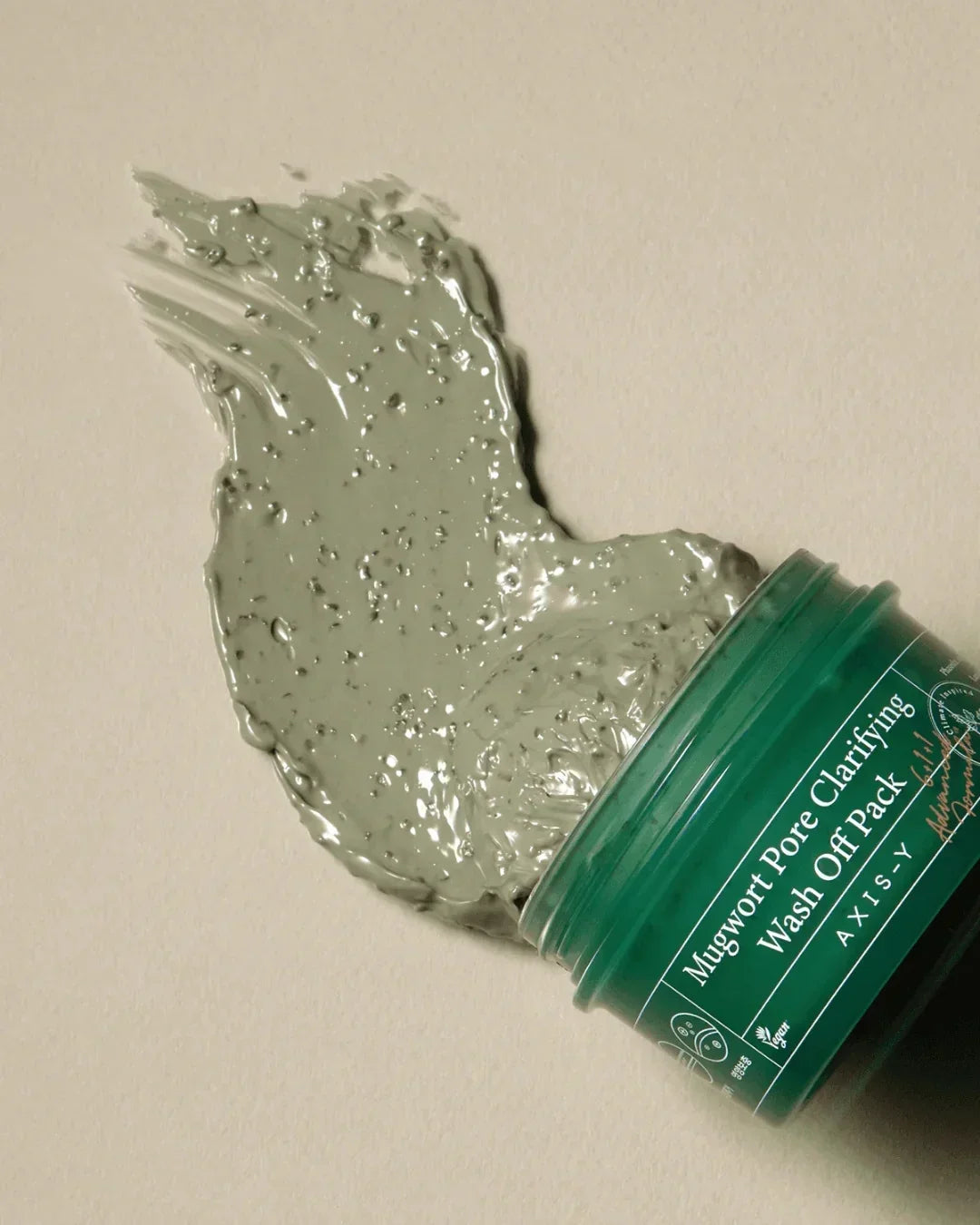 Opened green jar of Mugwort Pore Clarifying Wash Off Pack with product spread on beige background