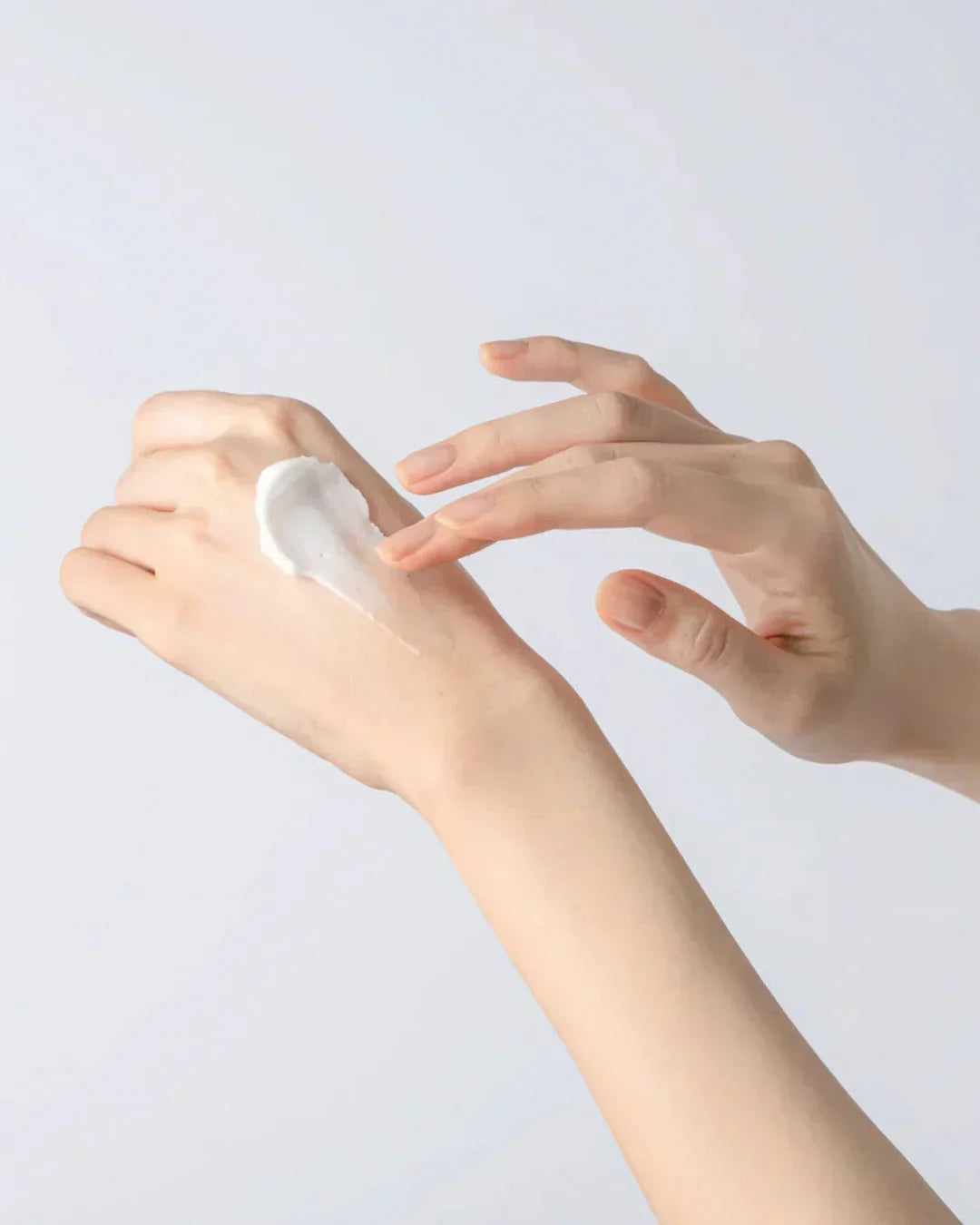Close-up of female hands applying creamy white moisturizer on back of hand against plain background