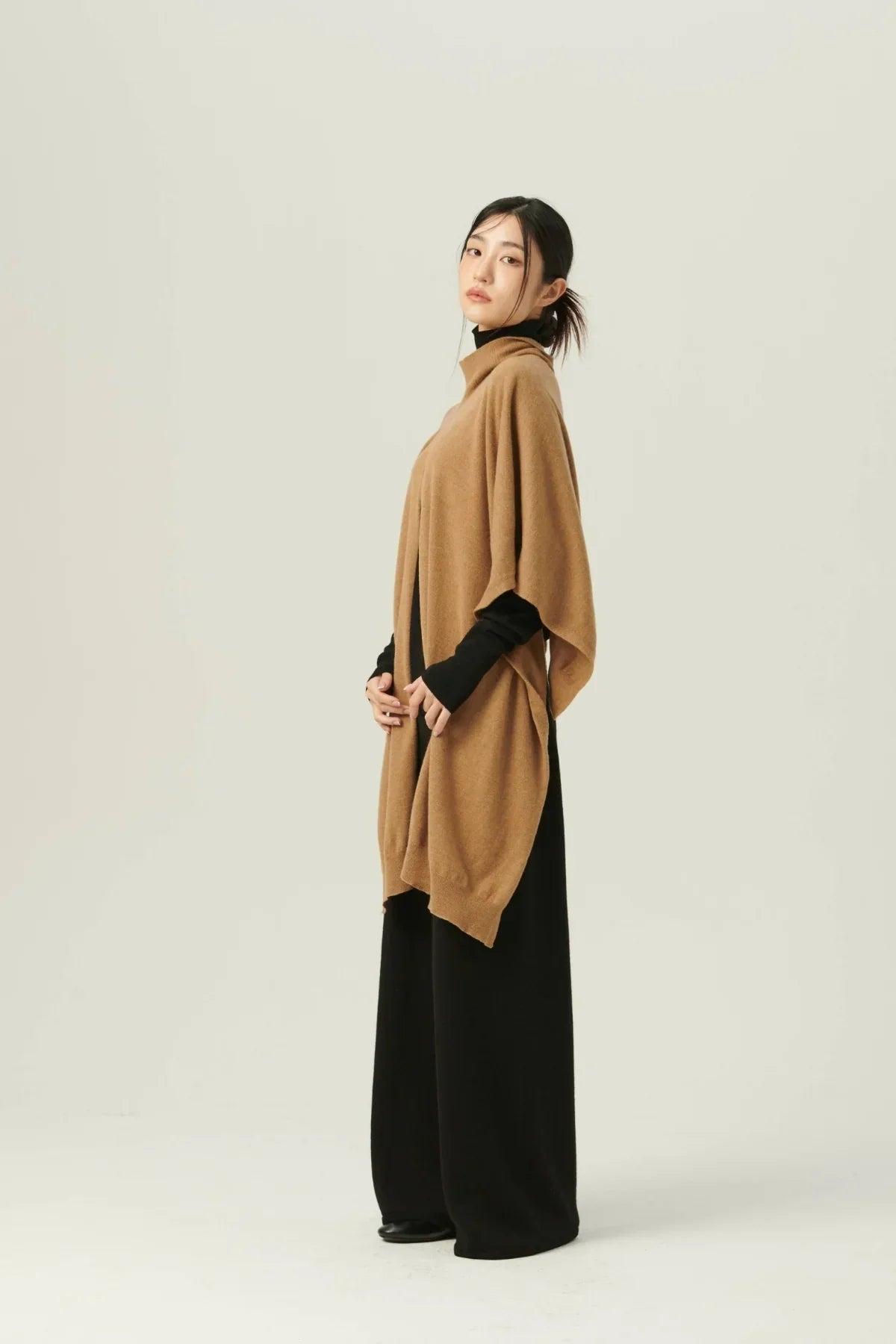 Woman wearing beige multiway knit cape over black turtleneck and wide-leg pants on neutral background