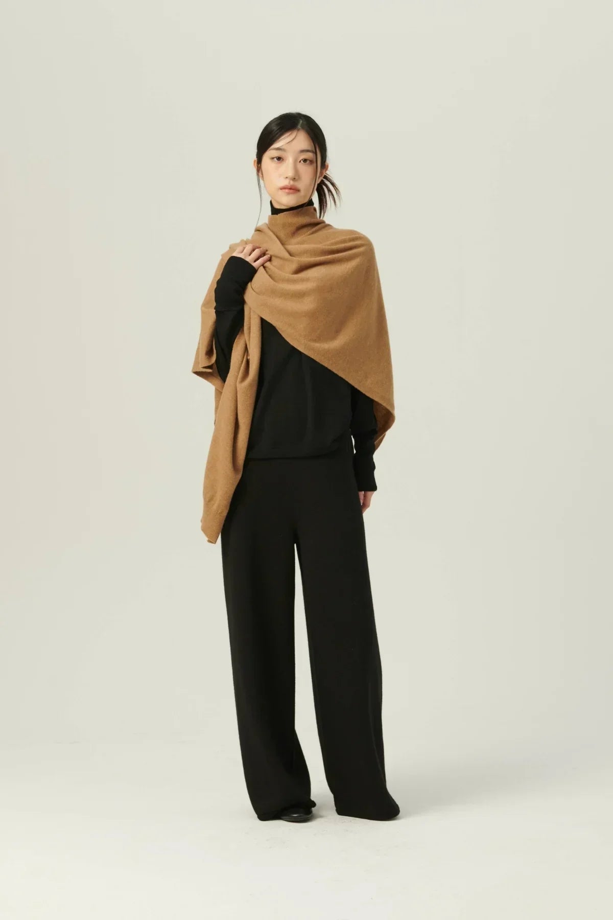 Woman wearing beige multiway knit cape over black outfit against plain background