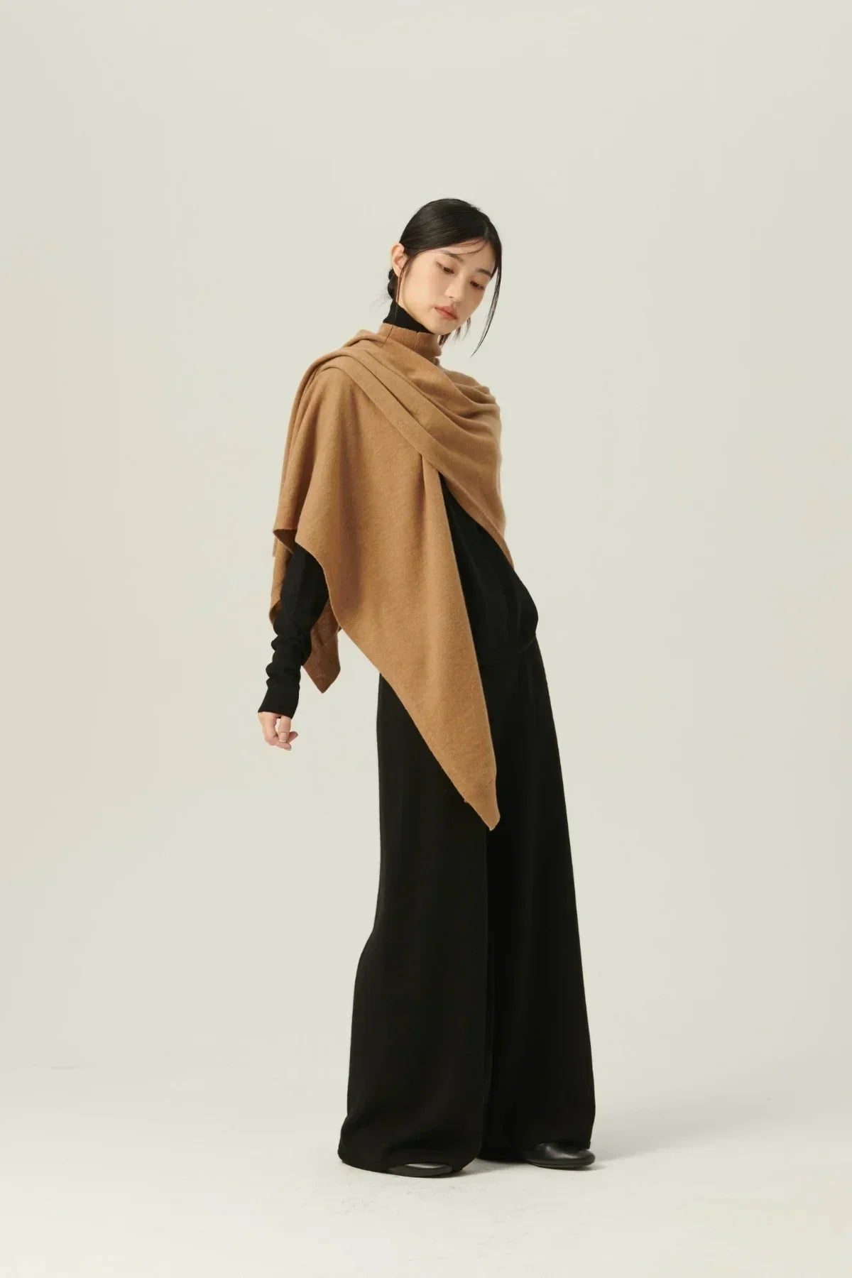 Model wearing beige multiway knit cape over black turtleneck and wide-leg pants on neutral background