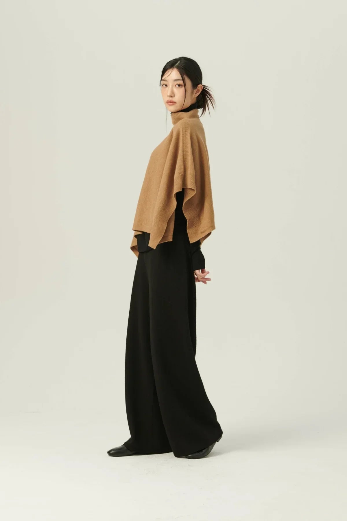 Woman wearing beige knit cape and wide-leg black pants in studio with plain background