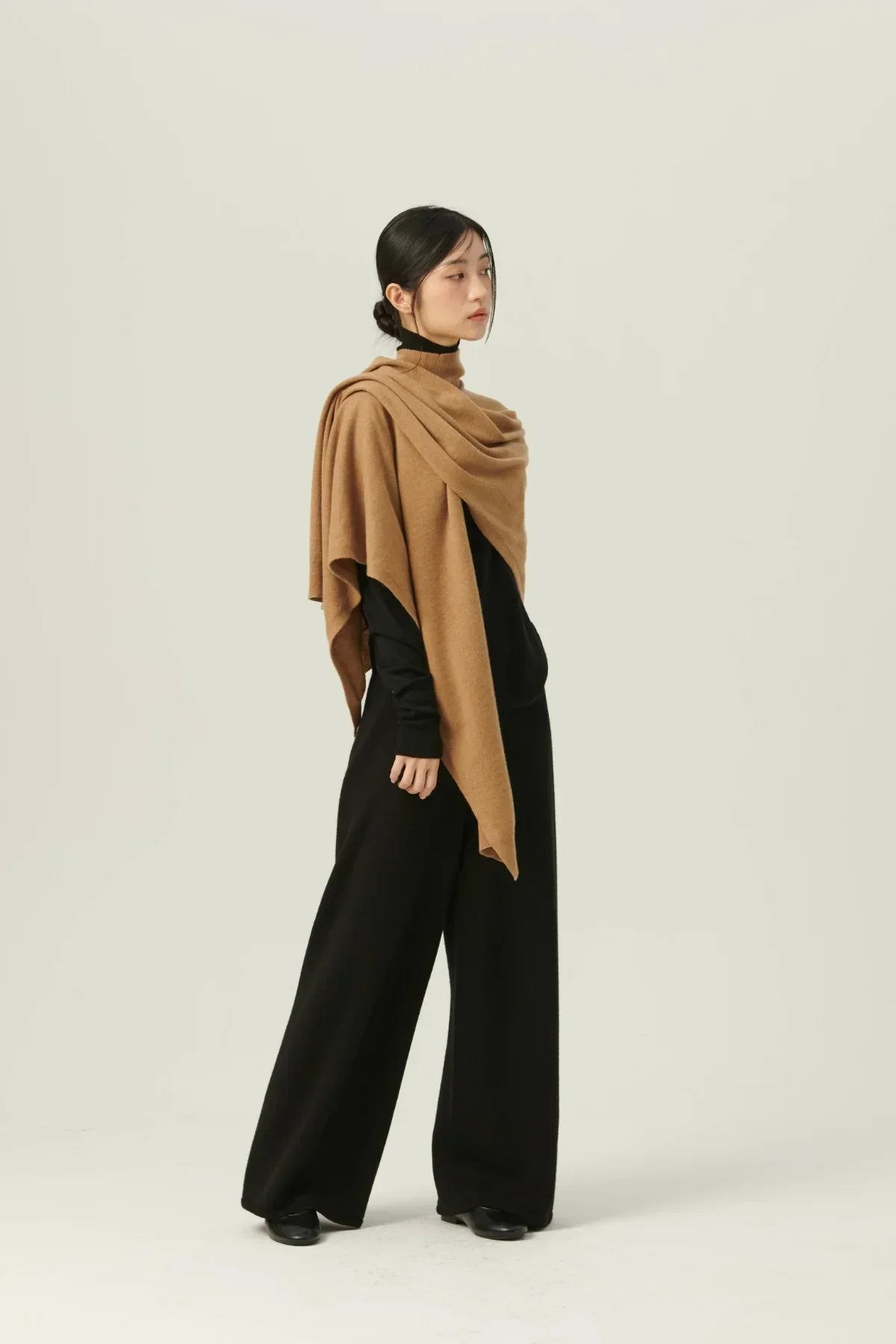 Woman wearing a beige knit multiway cape over black outfit in studio with plain background