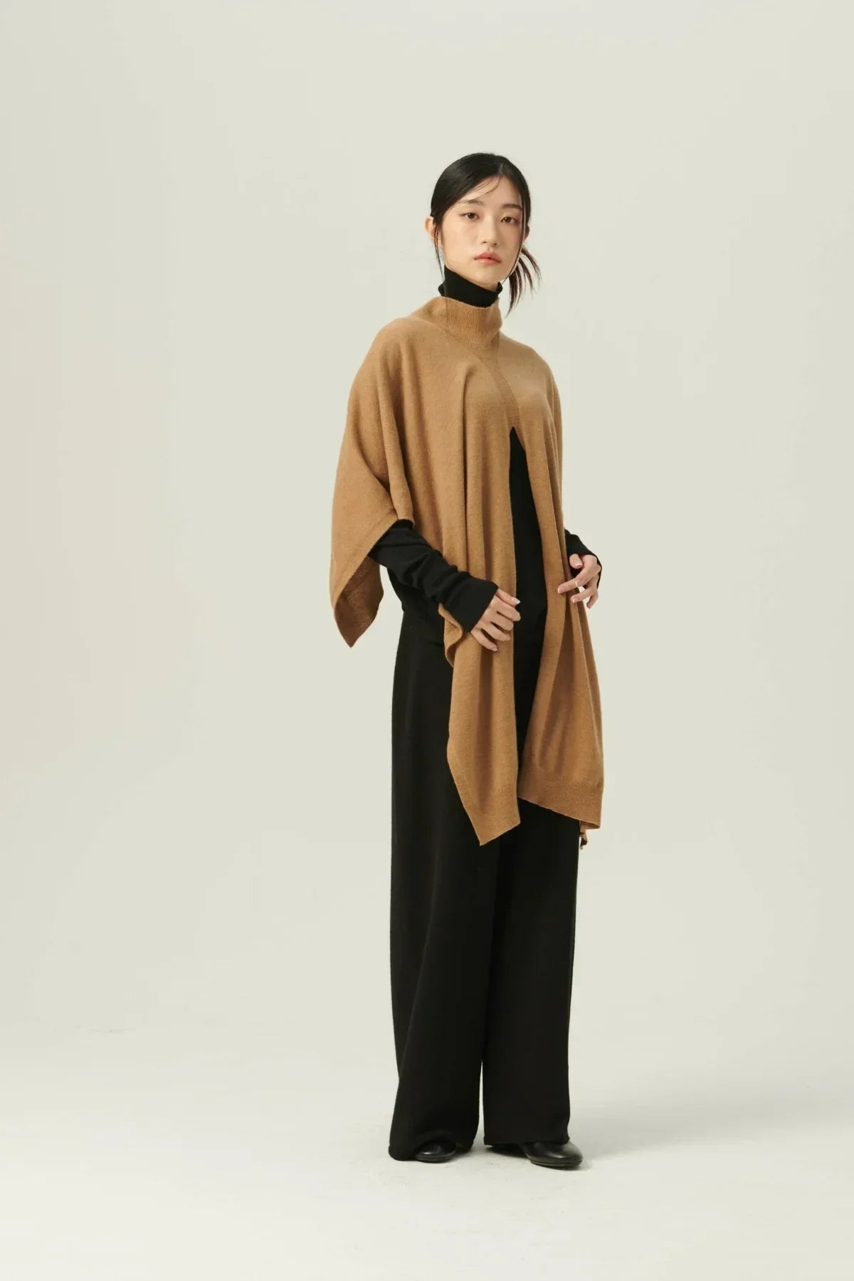 Woman wearing a beige multiway knit cape over a black turtleneck and wide-leg pants in a studio