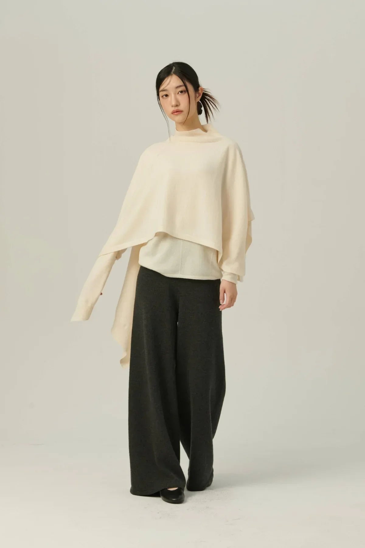 Model wearing ivory knit multiway cape with wide-leg black pants and black shoes on gray background