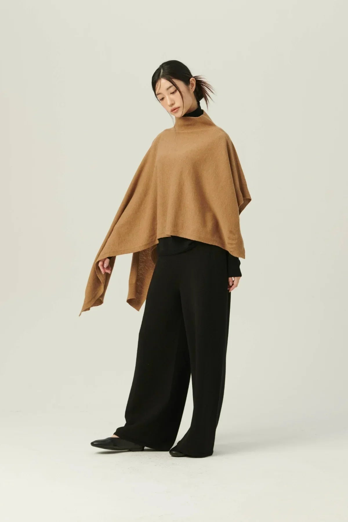 Woman wearing a camel-colored knit poncho with black wide-leg pants and black shoes against a plain background