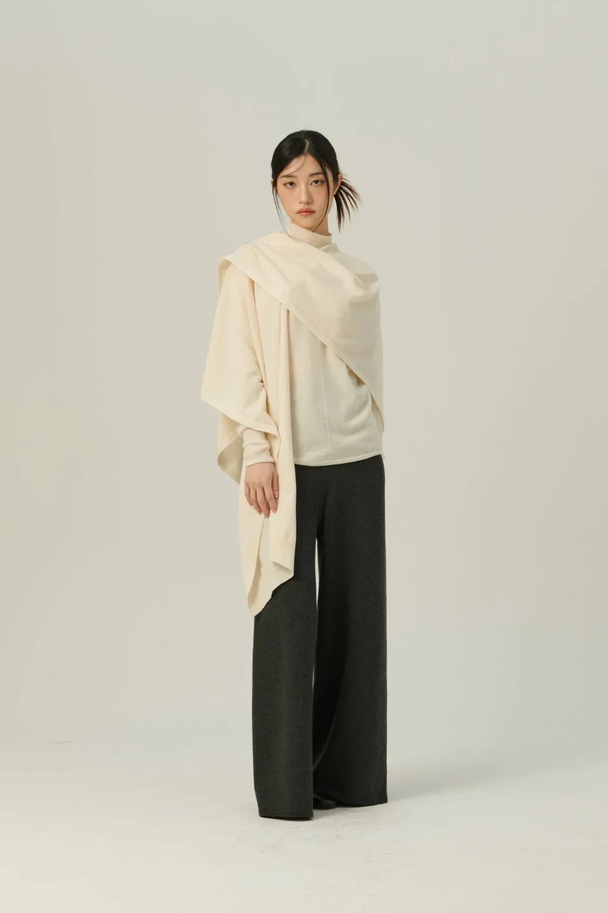 Woman wearing ivory knit multiway cape with dark wide-leg pants in minimalist studio